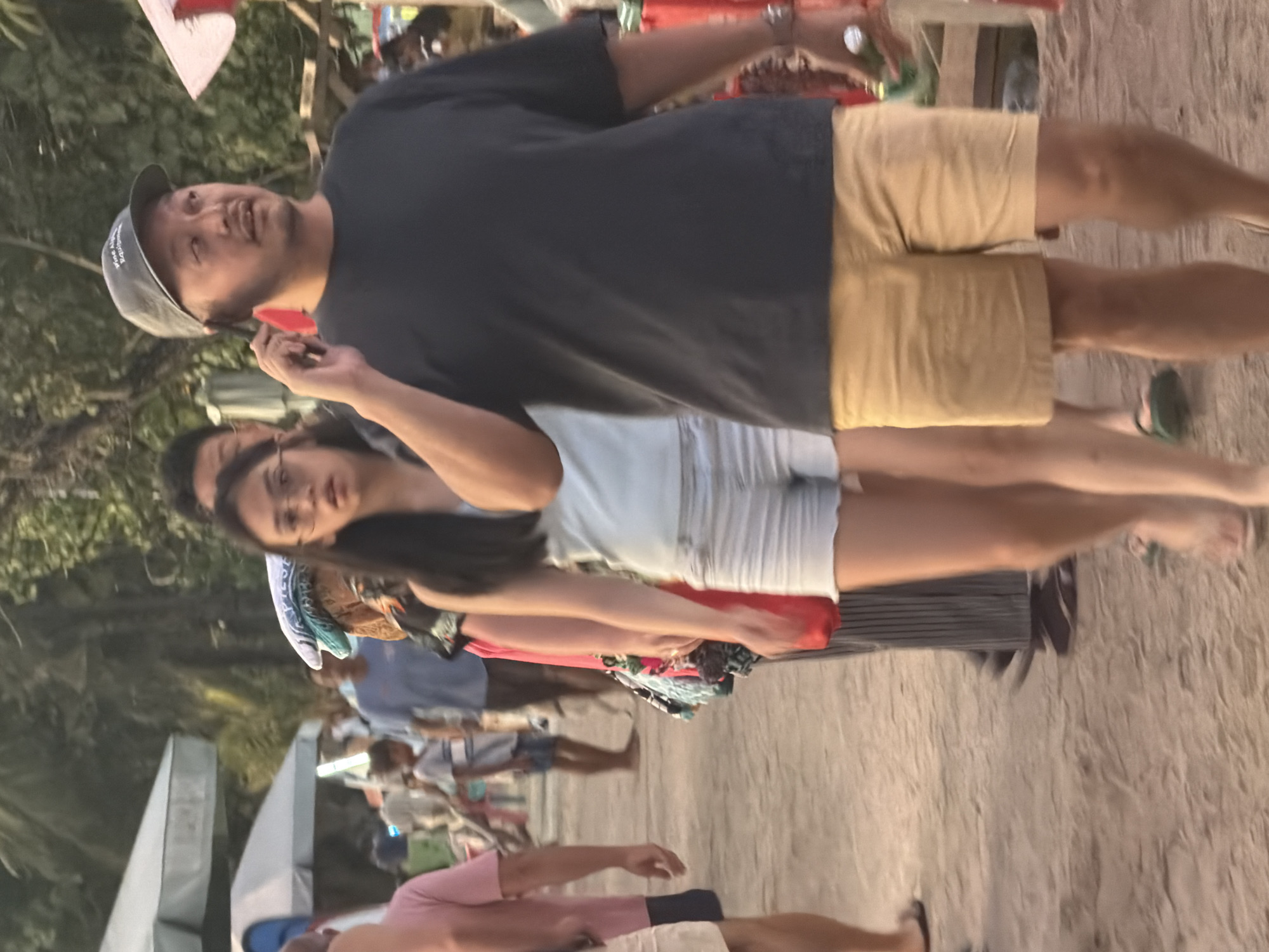 The image captures a street scene in Kuta, Indonesia, likely a marketplace or a bustling pedestrian area given the presence of stalls and people. The lighting suggests it is either early morning or late afternoon. In the foreground, a man wearing a dark t-shirt and light brown shorts is walking and talking on a cell phone, holding it to his left ear. He is wearing a grey baseball cap. To his left and slightly behind him, a young woman in a light blue top and white shorts is walking, also looking towards the left of the frame. Further in the background, other individuals are visible, including a person in a pink shirt on the far left, and several others further back near market stalls. The ground appears to be sandy or dirt. The overall atmosphere is casual and appears to be a public area with commercial activity.