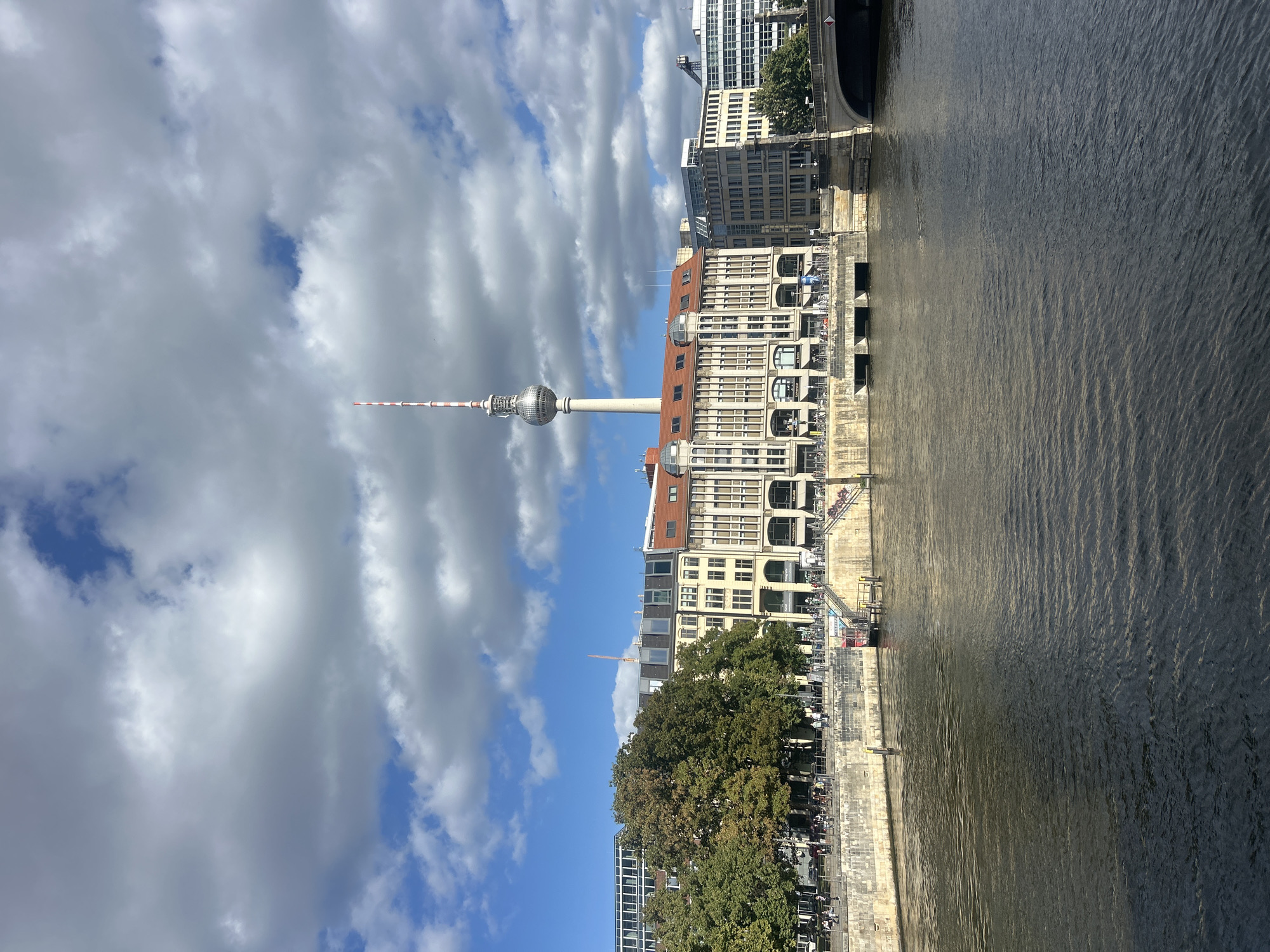 This is a photograph taken from a body of water, likely the Spree River, looking towards a waterfront cityscape in Berlin, Germany. The iconic Fernsehturm (Television Tower) is prominently visible in the background, its spherical observation deck and pointed antenna piercing the cloudy sky.
In the midground, a row of buildings lines the riverbank. These buildings are varied in architectural style, with some featuring red tiled roofs and others displaying more modern glass facades. A pedestrian walkway runs along the river, with people visible strolling and cycling. Bicycles are also parked along the embankment. Several trees, in full foliage, add greenery to the scene. Steps lead down from the walkway to the water's edge, and a small red structure, possibly a pier or boat dock, is visible.
The foreground is dominated by the dark, rippling water of the river, reflecting the sky and buildings. A bridge with arches spans the river in the far right of the image. The sky is a mix of bright blue patches and large, fluffy white and grey clouds. The overall impression is a sunny day with some cloud cover, typical of an urban European setting.