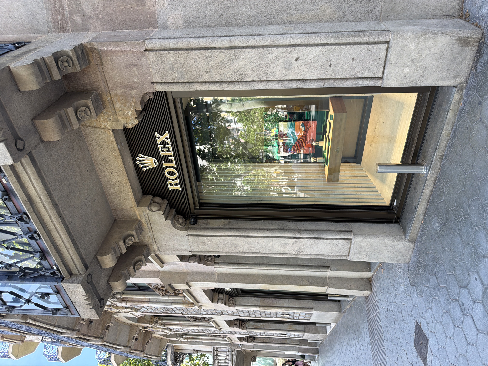 The image shows the facade of a Rolex store on a city street. The building is made of stone with ornate architectural details, including balconies with wrought-iron railings, decorative carvings, and fluted columns. The Rolex store has a large display window showcasing watches and a colorful artwork. The street in front of the store is paved with hexagonal tiles. A few people are visible walking in the background on the left side of the image. The scene appears to be in Barcelona, Spain, given the architectural style and the presence of palm trees reflected in the store window.