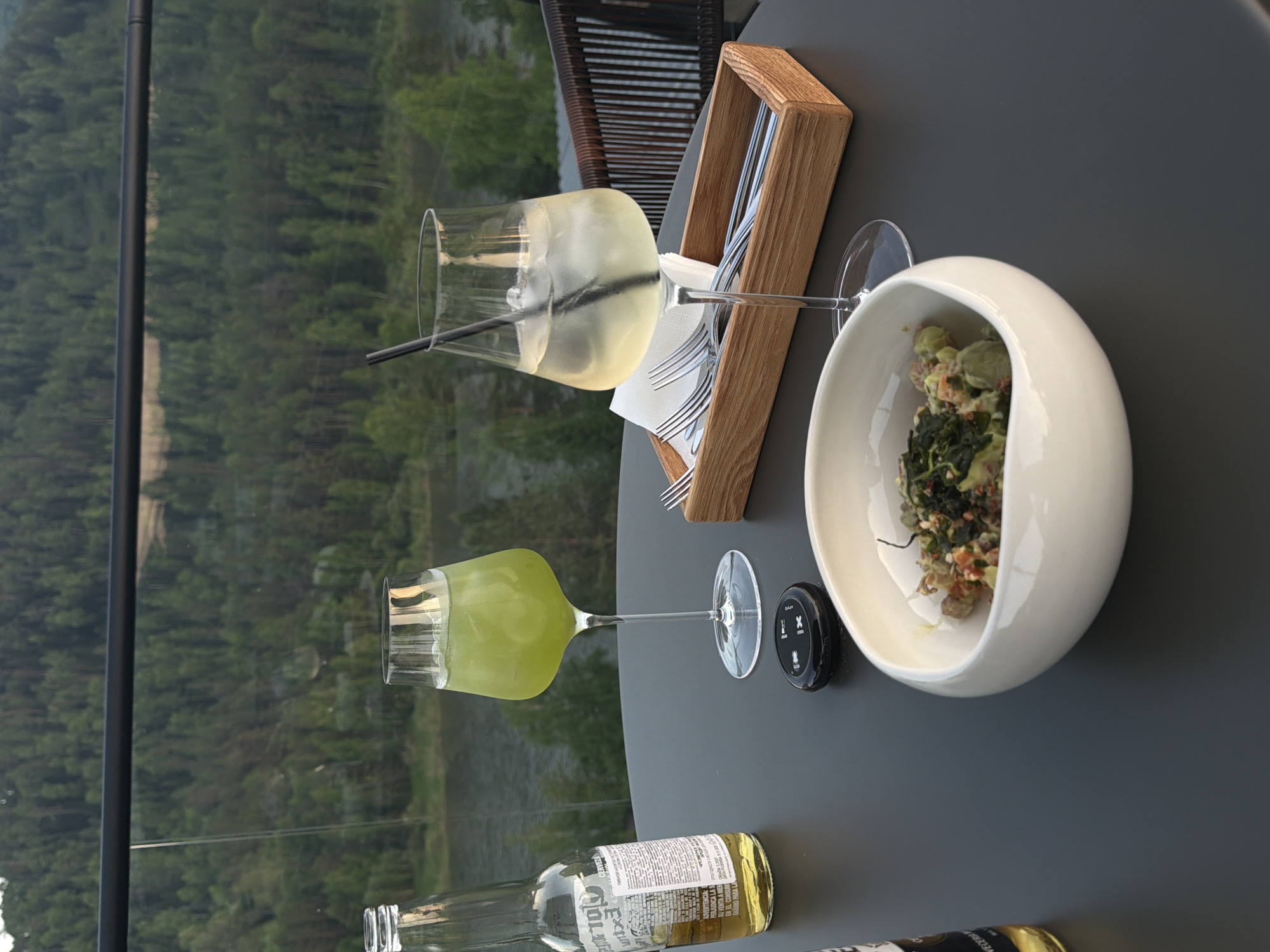 The image captures an outdoor dining scene on a terrace or balcony, overlooking a natural landscape in Манский, Russia. A dark grey, circular tabletop occupies the foreground, displaying several items. On the left, an empty Corona Extra beer bottle stands upright. Adjacent to it, a tall, stemmed glass contains a vibrant green iced beverage. A small, black, oval-shaped car key fob with white "iX" branding is positioned near the center. To its right, another tall, stemmed glass holds a pale yellow or clear iced drink with a black straw. In the immediate foreground, a white, irregularly shaped ceramic bowl contains a dish featuring diced green components (e.g., avocado), reddish elements, and dark green leafy ingredients. Behind the rightmost glass, a light wooden rectangular caddy holds several silver metal forks and white folded napkins. A dark brown woven outdoor chair back is partially visible on the far right. Beyond the table, a clear glass safety railing with a black top bar offers an unobstructed view of a wide, calm river with greenish-blue water flowing through the midground. The background consists of densely forested hills and mountains, under a softly lit sky, indicative of late afternoon or early evening.