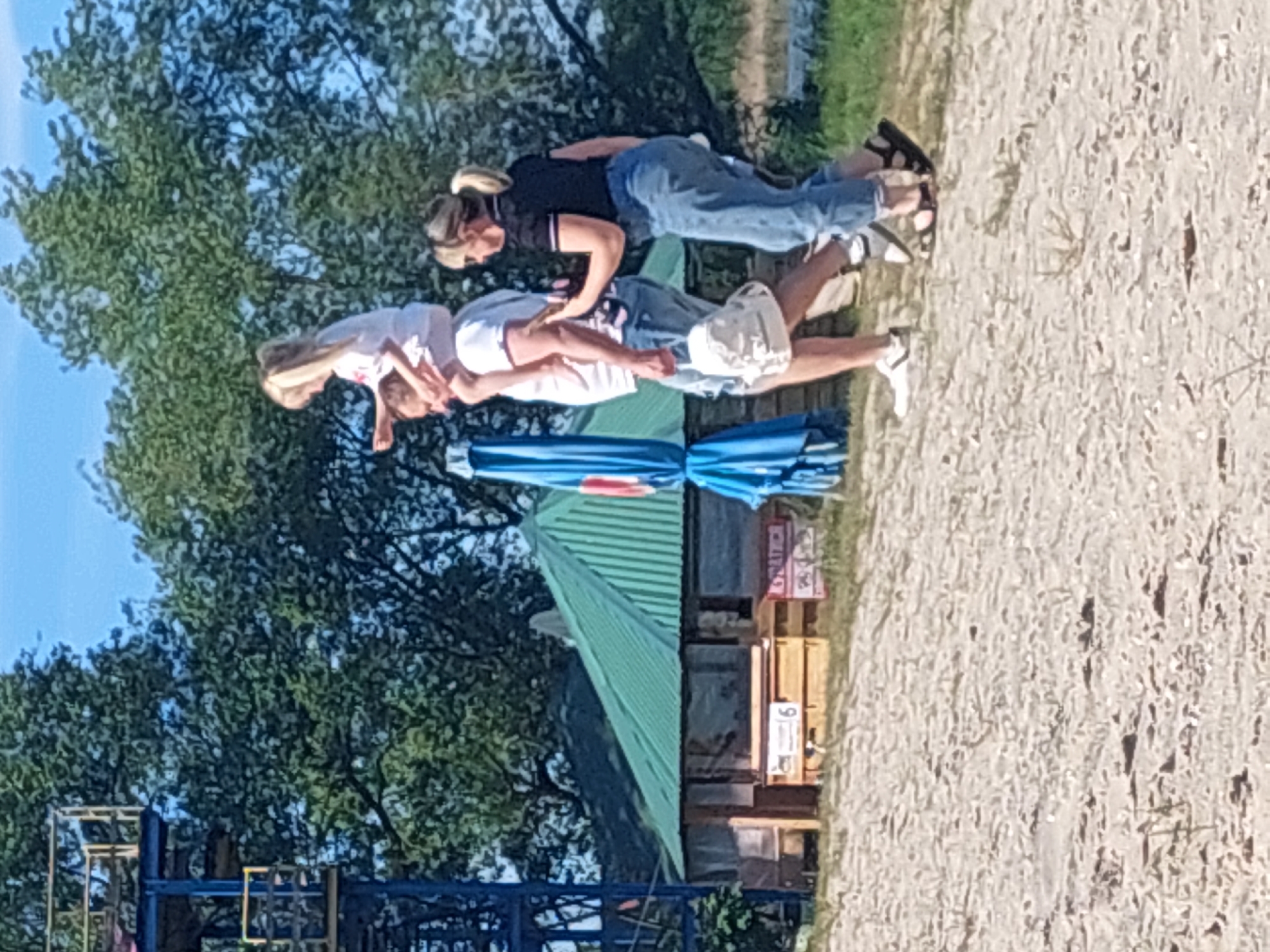 A medium shot displays three individuals horizontally stacked on top of each other in an outdoor setting, possibly a park or recreation area, with a sandy surface visible on the right. The bottom person is an adult male wearing light-colored shorts and a white shirt. The middle person is an adult female wearing jeans and a dark top. The top person appears to be a young child in a light-colored outfit. All three are facing the same direction. To the left of the figures, a blue and green structure is visible, suggesting a possible playground or recreational facility. In the background are trees and some indeterminate buildings. The image lacks any specific markers to definitively locate the scene within the city of Ternopil, Ukraine. The action is people lying on top of each other.