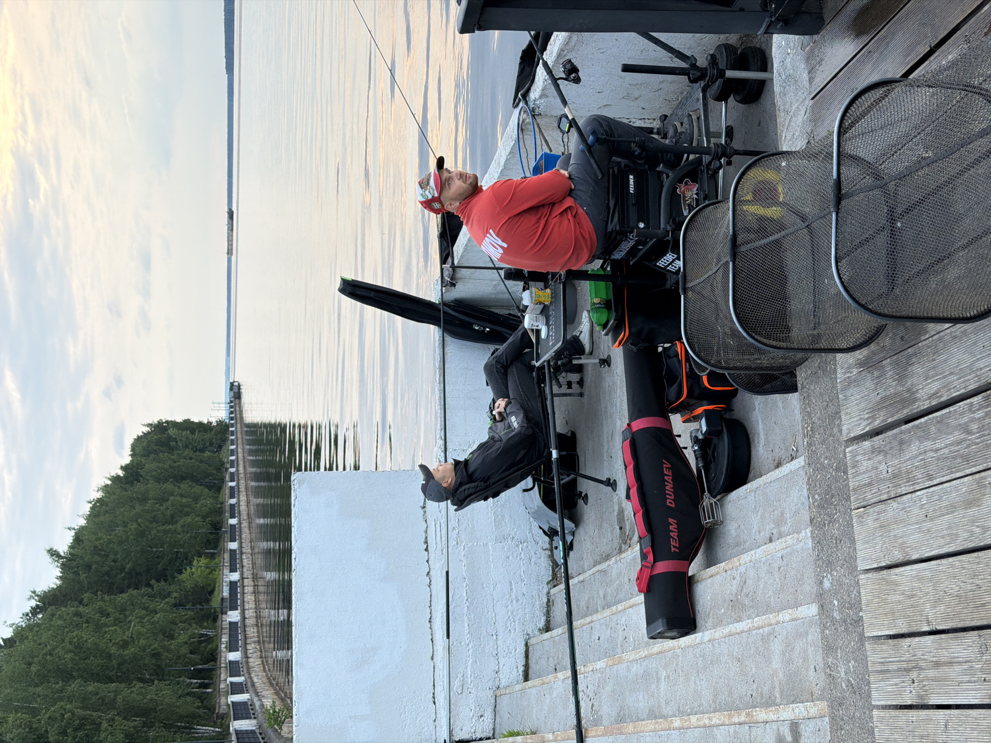 This image captures an outdoor fishing scene along a large body of water in Chaikovsky, Russia, likely on the Kama River. The scene depicts two men engaged in feeder fishing from a concrete embankment that transitions into a wooden deck.
The foreground features a man seated on a sophisticated fishing chair with multiple attachments, including rod rests and bait trays. He is wearing a red long-sleeved shirt with "МОИ" (MOI) visible in white letters on the back, dark pants, and a baseball cap. His gaze is directed upwards and towards the water, suggesting observation or anticipation. Near him, another man, dressed in a dark jacket, dark pants, and a baseball cap, is also seated on a similar fishing chair setup, positioned slightly lower on concrete steps. Both men have multiple fishing rods deployed into the calm water.
Surrounding the fishermen are various pieces of specialized angling equipment. A prominent black and red fishing bag, clearly labeled "TEAM DUNAEV" in white text, rests on the concrete steps. Two large, rectangular mesh keepnets are positioned on the wooden deck, ready for fish. Other visible details include "FEEDER TEAM" text on some of the equipment boxes, additional bait boxes, and fishing tackle.
The setting includes a calm body of water reflecting the sky, which appears partly cloudy, indicating either early morning or late afternoon. Across the water, a distant, tree-lined shore is visible, along with a long, low linear structure, possibly a dam or a bridge, which appears to have a conveyor system. The overall atmosphere is one of calm concentration, typical of a dedicated fishing session or competition.