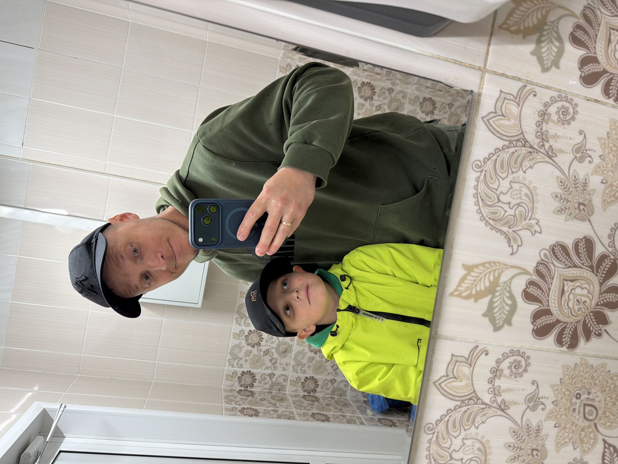 The image captures a mirror selfie featuring an adult male and a child male in what appears to be a public or private restroom in Birobidzhan, Russia.
The adult male, appearing to be in his 30s to early 40s, is positioned on the right side of the frame. He has light stubble, blue-green eyes, and wears a black baseball cap with a dark "NY" logo and an olive green hooded sweatshirt. A gold-toned ring is visible on the ring finger of his left hand. He holds a dark-colored smartphone, equipped with three visible camera lenses, in his left hand, taking the photograph. He gazes directly towards the phone's lens with a slight, neutral expression.
To the adult's left and positioned lower in the frame is a child male, estimated to be between 6 and 8 years old. He wears a black baseball cap featuring an orange or brown "M" or "W" logo and a bright neon yellow waterproof jacket with a visible green inner lining at the collar. His blue eyes are directed upwards and to his left, away from the camera, giving him a contemplative expression. The child appears unengaged with the photo-taking process.
The background consists of tiled walls. The upper section features plain, light beige rectangular tiles, while the lower section is adorned with intricate floral patterns in shades of beige, brown, and subtle green. A white door frame with a visible door closer mechanism is on the far left. A blue plastic trash bag is partially visible inside a container near the child's left arm. A white, rectangular cabinet or shelf is reflected in the upper right portion of the mirror, and a portion of a white wall-mounted fixture is visible on the far right.