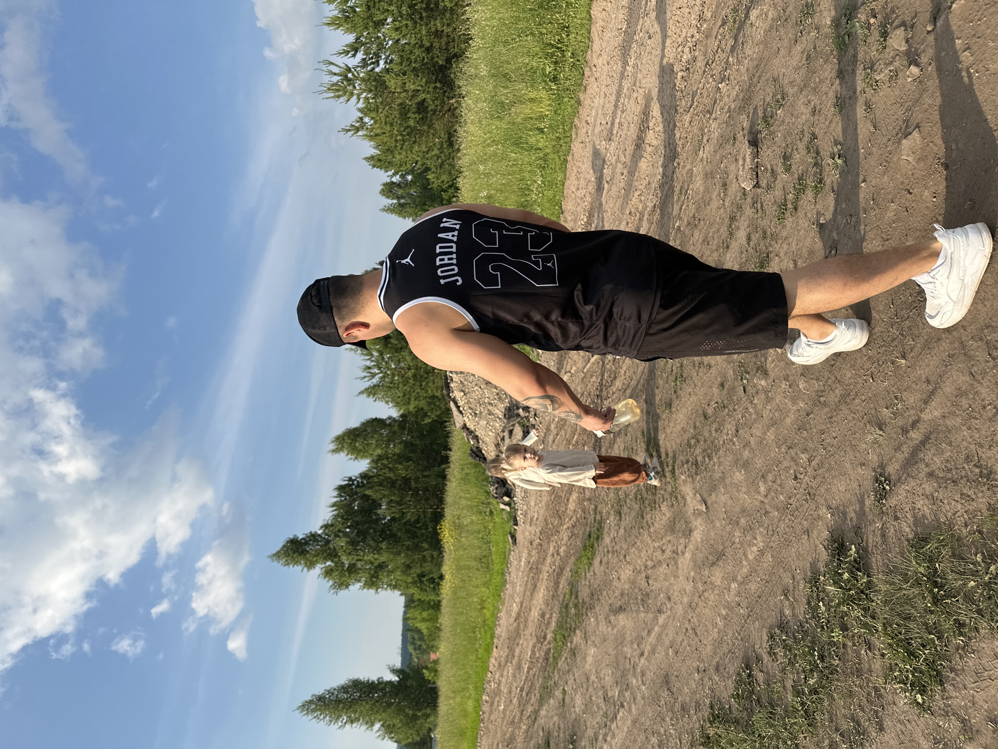 In a rural setting with a clear blue sky and scattered clouds, a man wearing a black Jordan jersey with the number 23 and black shorts walks away from the camera on a dirt path. He is holding a clear plastic bottle containing a yellow liquid. To his left, a young child in a light-colored shirt and brown pants stands looking towards the camera. The path is flanked by green grass and trees, with a pile of debris visible in the background. The scene appears to be on the outskirts of Эстачи, Russia.