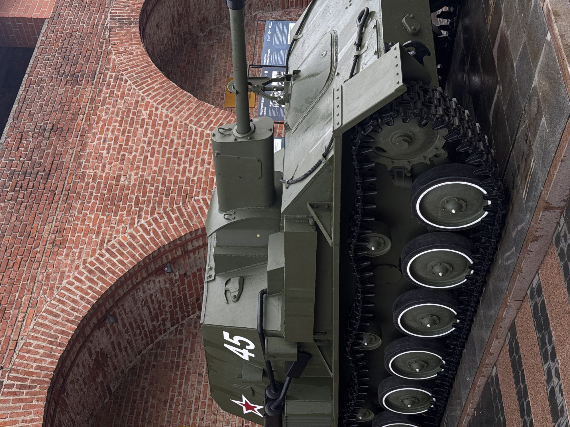 The image shows a Soviet T-34-85 tank, a medium tank used in World War II, on display as a monument. The tank is painted in a dark green color with the number "45" and a red star painted on the front. It is positioned next to a red brick building with an arched entrance. The location is likely a memorial or park in Nizhny Novgorod, Russia, given the type of tank and the architectural style of the building. The tank's wheels are black with white circles painted on them, and the treads are black. The monument appears to be partially set into a granite base, which is seen on the right of the frame.