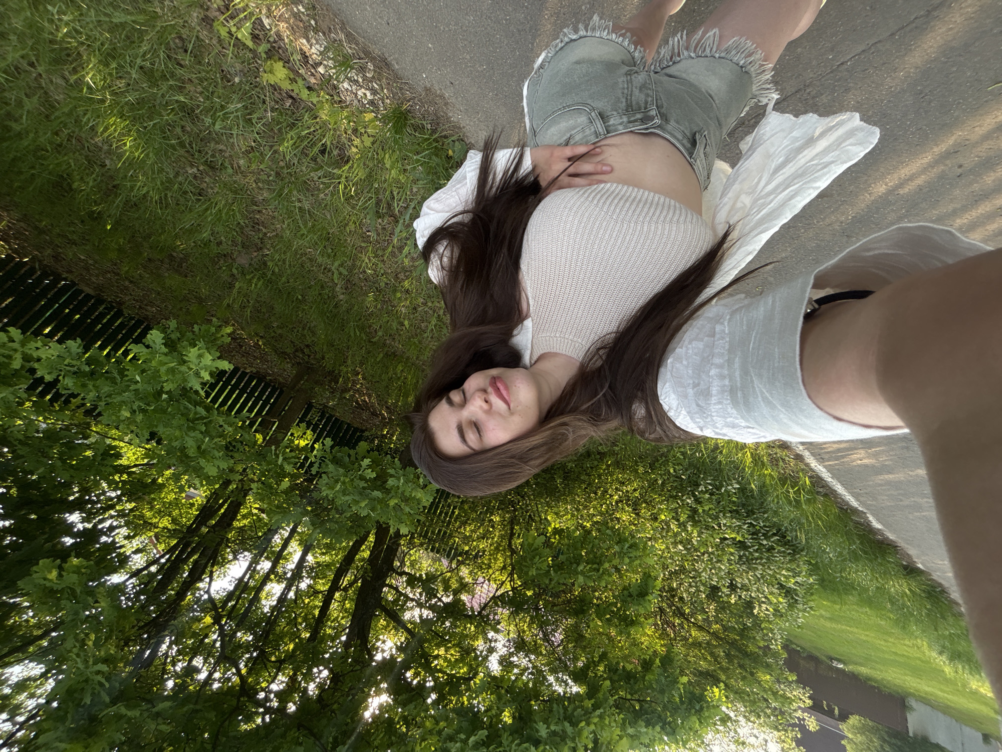 A woman is lying on her back on a paved surface near a grassy area, possibly a sidewalk or pathway in a park. She is wearing a light-colored knitted top, denim shorts with a frayed hem, and a white shirt. Her eyes are closed. Trees with dense foliage are overhead, filtering sunlight. The location appears to be a green area within a city. It is not possible to definitively confirm the specific location of Мещёры, Russia, based on the provided image.