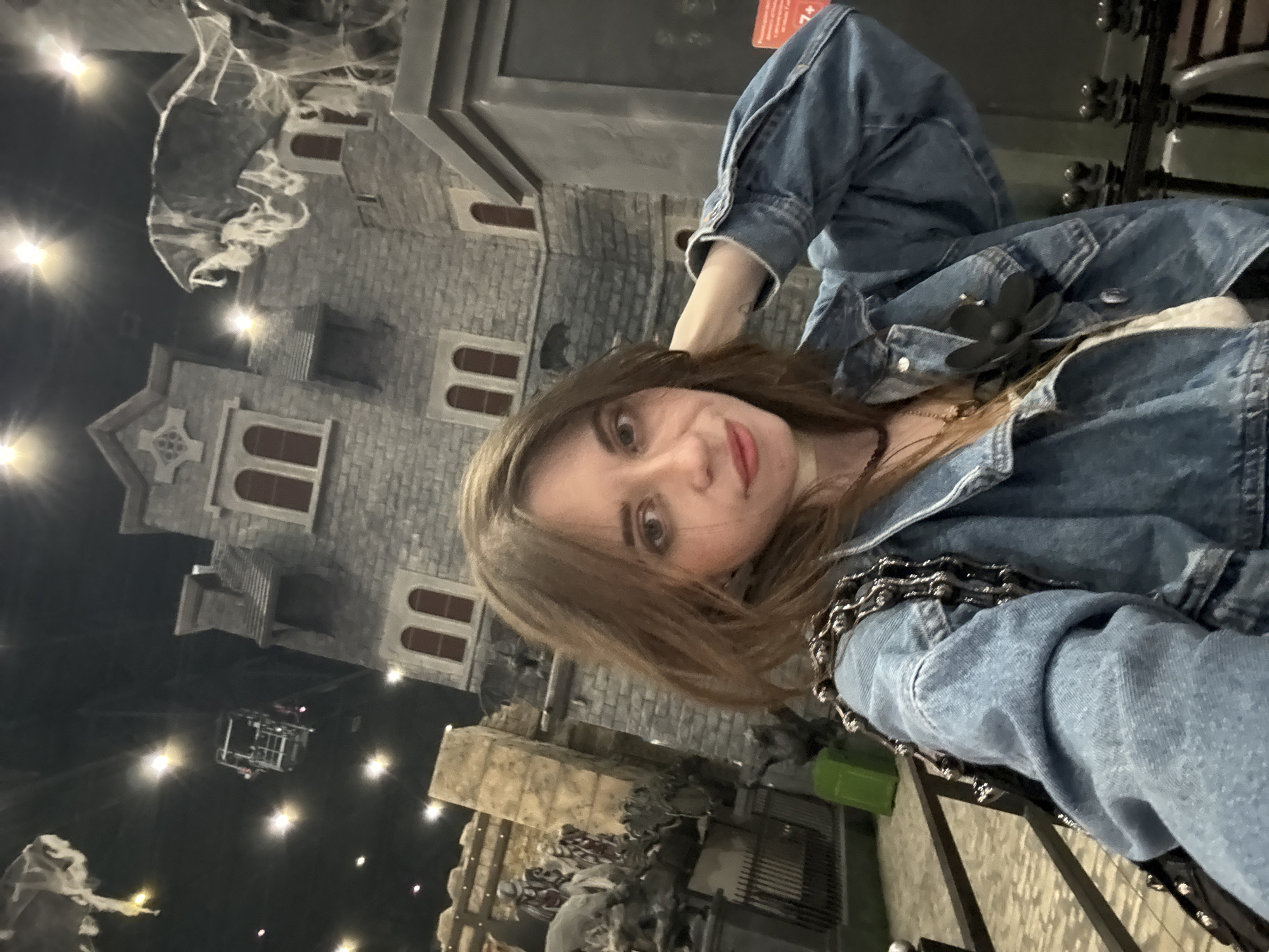 The image is a low-angle selfie of a fair-skinned woman with light brown hair, wearing a denim jacket. She appears to be indoors at a themed location, likely a Halloween or horror-themed attraction. In the background, there is a large grey structure resembling a haunted house or castle with multiple floors and windows with red frames. There are theatrical fog or cobwebs draped on the structure. In the lower portion of the frame, there are additional figures or statues that are indistinct due to the low light conditions. A metal railing is visible in the foreground. The environment seems dimly lit with spot lights directed towards the set pieces. The location is possibly in Moscow, Russia.