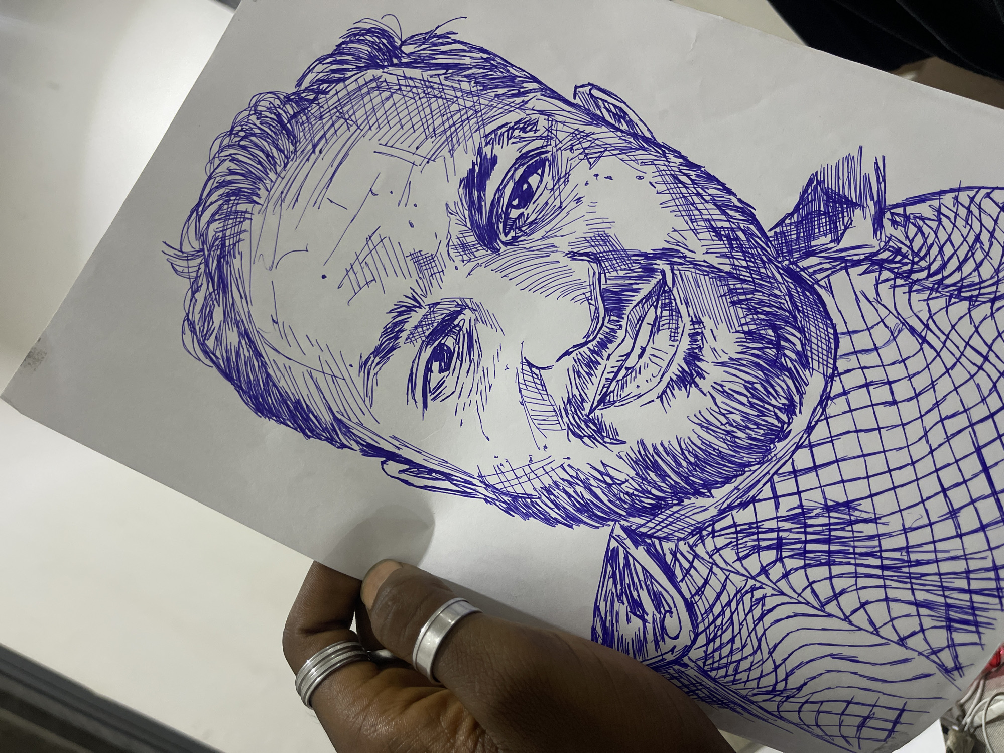 A close-up, overhead shot captures a hand holding a sketch of a man's face. The sketch is done in blue ink on white paper, featuring cross-hatching to create shading and texture. The man in the sketch has a beard, receding hairline, and is smiling. The hand holding the paper is dark-skinned and wears two silver rings on its index finger. The background is a plain white surface, possibly a table or desk. The image is shot in natural lighting conditions. The overall composition is focused on the sketch and the hand holding it.