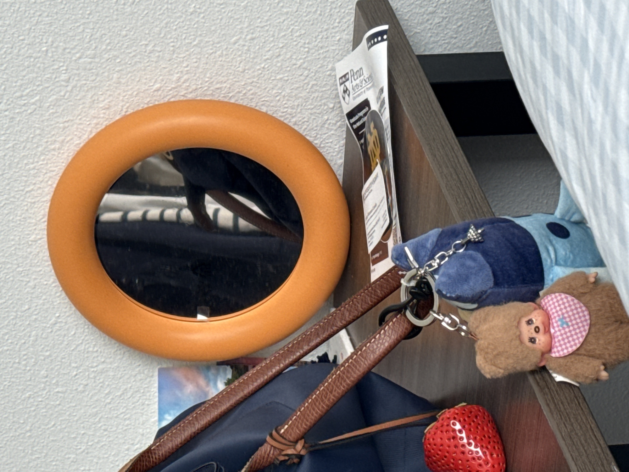 An indoor scene displays a collection of personal items against a white textured wall and wooden furniture, likely a shelf or bunk bed frame.
The central subject is a circular orange-framed mirror, reflecting a person's torso and arm wearing dark clothing. Below and to the right of the mirror, a dark blue bag or backpack with brown leather straps is visible. One of the leather straps features intricate stitching and is adorned with a red, strawberry-shaped object, possibly a decorative pouch or charm.
Attached to the bag strap by a metal chain and keyring are two plush keychains. One is a dark blue, somewhat amorphous character, while the other is a brown monkey-like doll wearing a pink and white checkered bib.
To the right of the mirror, tucked between the wall and the wooden furniture, is a white piece of paper or flyer. Visible text on the flyer includes "Penn Arts & Scien" and "University of Pe", strongly indicating "University of Pennsylvania Arts and Sciences." Other smaller text and logos are present but illegible.
The lighting suggests it is daytime, providing a clear view of the textured wall and the various objects. No explicit activity is occurring; the items are stationary. The setting appears to be a personal space, possibly a student's room.
Location context: Austin, United States of America.