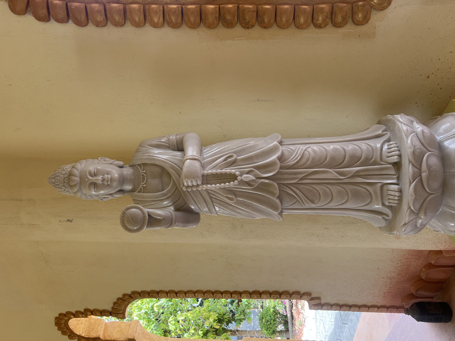 A full-body standing statue, crafted from light-colored stone, likely marble, is centrally positioned against a smooth, light yellow wall in the city of Đăk Mar, Vietnam. The statue depicts a serene Buddhist deity, possibly Guanyin, adorned with an ornate crown, flowing robes, a beaded necklace, and bracelets. Its raised right hand holds a disc prominently featuring the Yin-Yang symbol. The left hand holds a string of white prayer beads (mala) with a tassel, draped downwards. The statue rests on an elaborate lotus-shaped pedestal. To the immediate left, an arched opening framed by a reddish-brown textured border provides a view of an outdoor environment with lush green foliage and a paved ground. A dark, cylindrical object is situated on the ground near the base of this archway. A similar reddish-brown textured architectural element is partially visible along the far right edge of the frame.
