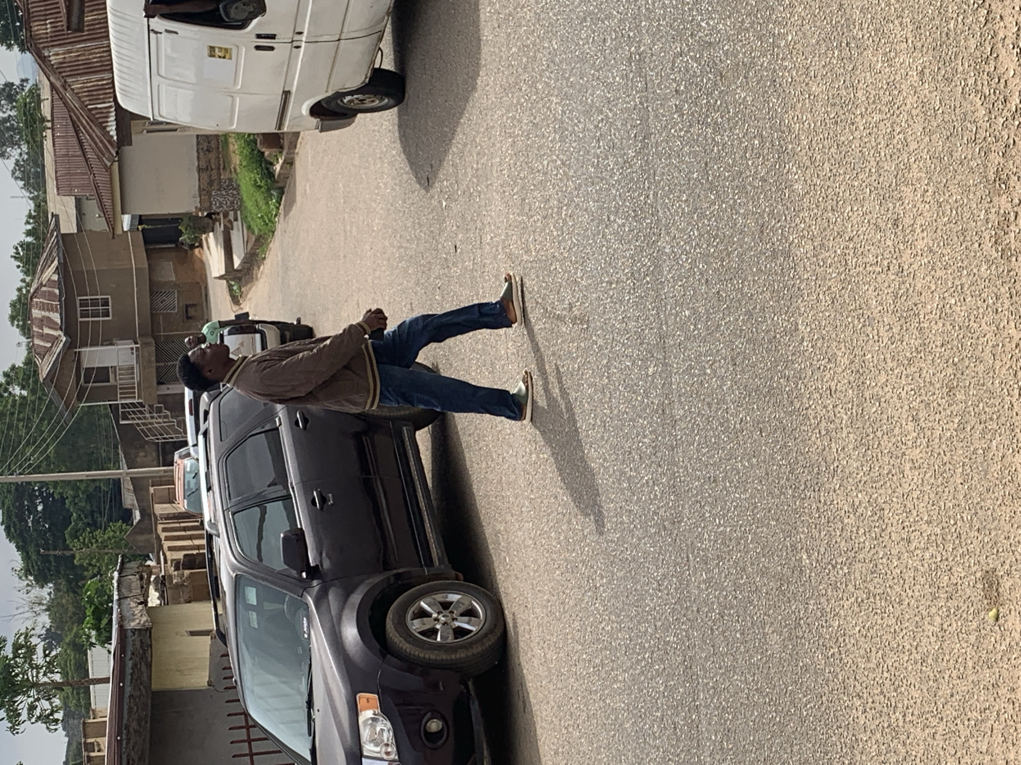 The image displays a sunlit street scene, likely in a residential or semi-urban area of Jos, Nigeria. A man stands in the mid-ground, turned slightly away from the camera, positioned next to a dark-colored SUV that is parked on the side of the road. He is dressed in a brown jacket, blue jeans, and light-colored slip-on shoes, with his hands clasped in front of him, suggesting he is waiting or engaged in a static activity like observing or conversing.
Further down the street, a white commercial van is also parked, with another person in a light green shirt partially visible near it. The road surface is paved, possibly asphalt, showing a granular texture. Strong sunlight illuminates the scene, casting sharp shadows from the vehicles and the man, indicating a bright, clear day, likely late morning or early afternoon.
In the background, several buildings with reddish-brown corrugated metal roofs and light-colored concrete walls line the street. Utility poles with power lines are visible on the left side of the frame, and some sparse vegetation and trees can be seen beyond the structures. The overall setting depicts an ordinary daily street environment, with no specific event or intense activity clearly taking place. A small, yellow sticker is present on the side of the white van, but its text is illegible.