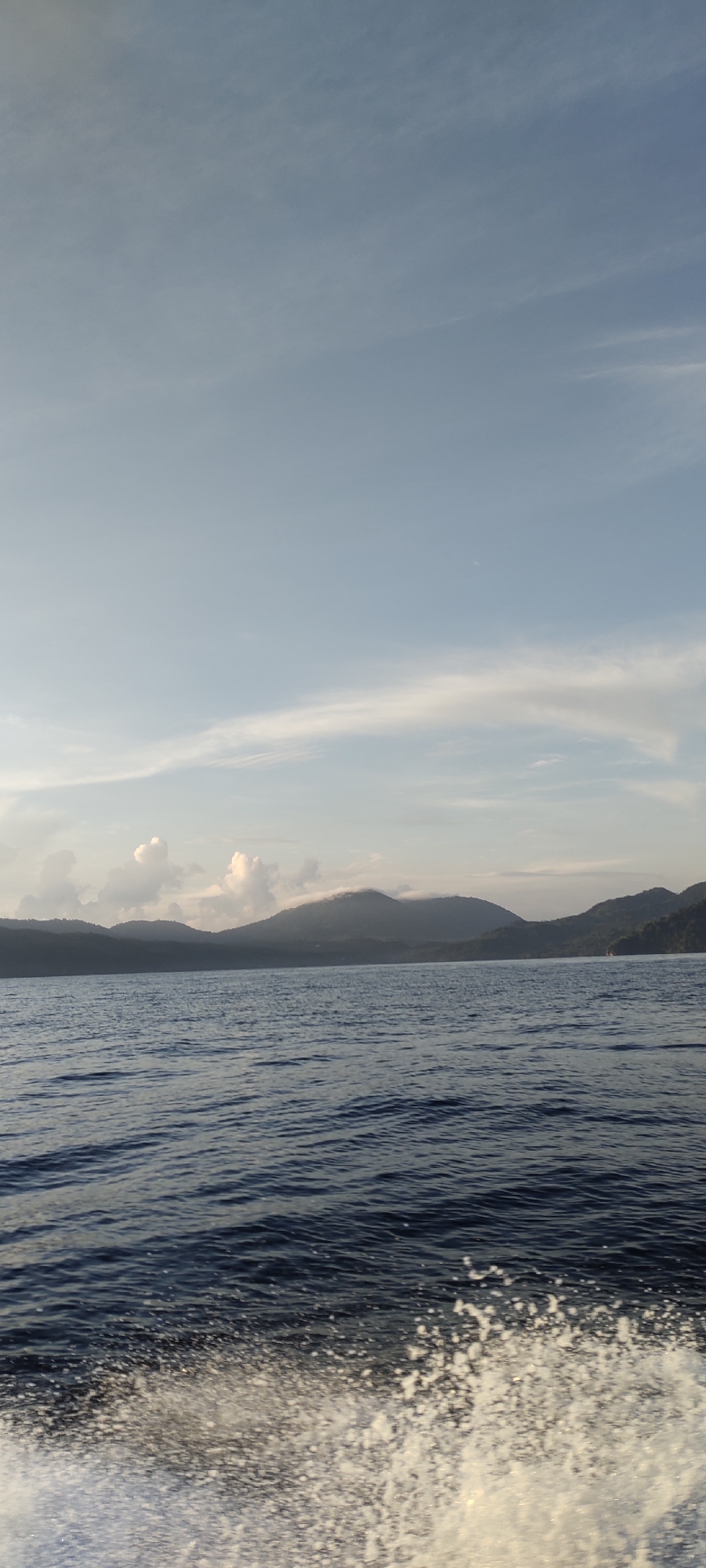 A serene seascape unfolds with a view of lush, mountainous islands in the distance, possibly located off the coast of Sabang, Indonesia. The water is a deep, calm blue, with gentle ripples across its surface, transitioning to a foamy white wake in the foreground, suggesting movement, likely from a boat. The sky above is a pale blue, adorned with wispy white clouds that add a soft texture to the horizon. The overall atmosphere is peaceful and natural, with the islands silhouetted against the sky.