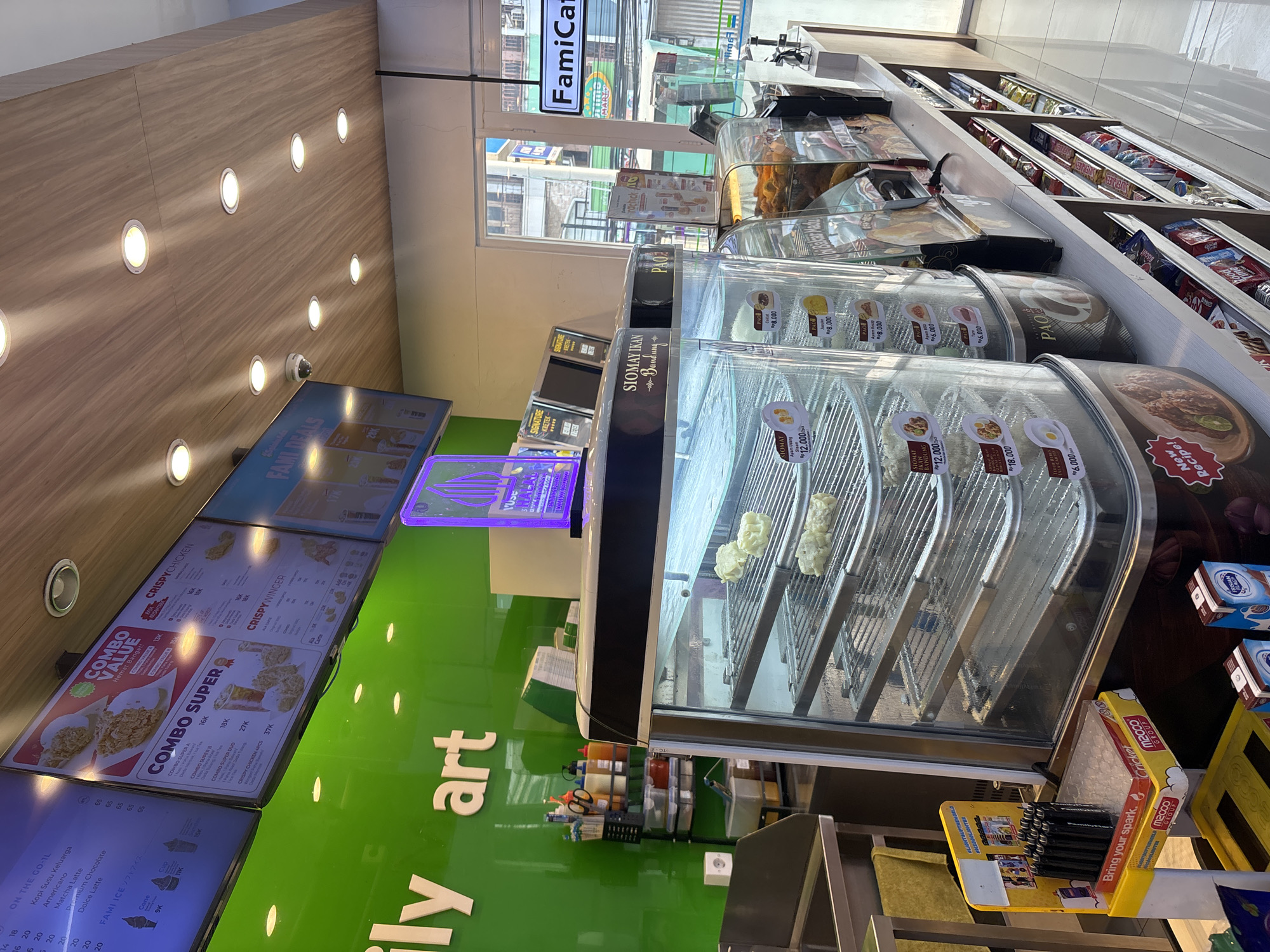 The image shows the interior of a FamiCafe convenience store, likely located in West Jakarta, Indonesia. The store features bright green walls and a wooden-paneled ceiling with recessed lighting. On the left, a large green wall displays white lettering that reads "ily art". Above this, a digital menu board displays various food and drink options, including "COMBO VALUE", "COMBO SUPER", and "CRISPY CHICKEN". In the center of the frame, a refrigerated display case showcases steamed dumplings, likely siomay, on metal racks. A vertical sign above the case reads "SIOMAY IKAN - Bandung". To the right, another display case contains fried food items and baked goods. Shelves along the right wall are stocked with various packaged snacks and beverages. A security camera is visible on the ceiling. The overall environment appears clean and well-lit, typical of a modern convenience store.