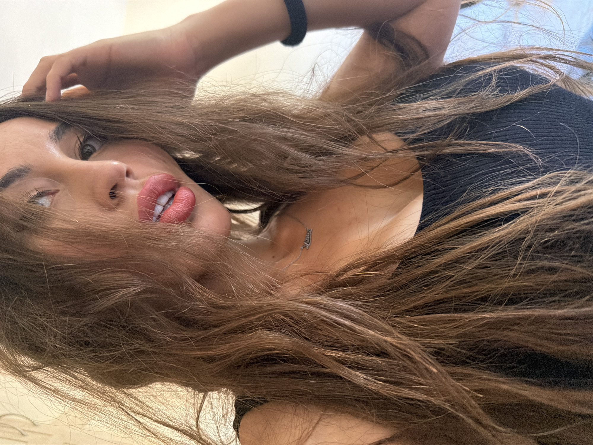 A medium close-up shot captures a woman with long, brown, wavy hair from the shoulders up. Her hair falls across her face, partially obscuring her features. She is wearing a black, sleeveless top and a silver necklace with the word "LOVE" inscribed on it. Her lips are painted a vibrant coral color, and her eyes are a striking blue-green. One hand is raised, fingers touching her hair near her temple. A dark band is visible on her right wrist. The background appears to be an indoor setting with a textured, beige wall on the left and a lighter, possibly blue-toned area on the right. The lighting suggests a bright, natural light source, casting shadows and highlighting strands of her hair. There is no indication of a specific location in Tersefanou, Cyprus.