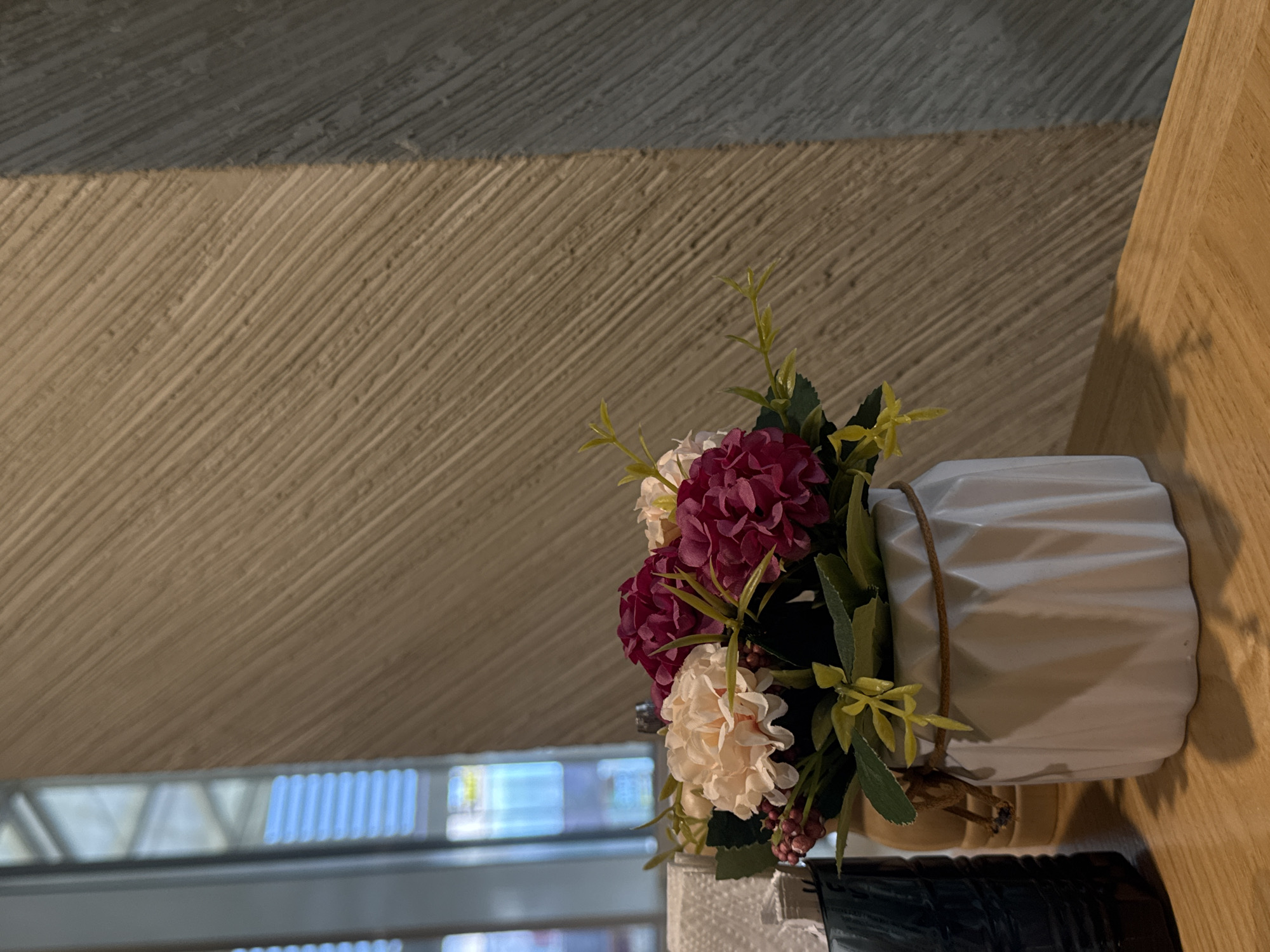 The image displays a still life composition featuring a vase of artificial flowers placed on a light wooden surface. The flowers consist of a cluster of rich burgundy blooms, complemented by lighter cream-colored flowers, all interspersed with green leaves and slender, light green stems. Some small, dark red berry-like clusters are also visible among the foliage.
The vase holding the flowers is white with a distinct textured, faceted, or ribbed design. A thin, brown, twine-like string is wrapped around the upper part of the vase. The wooden surface beneath the vase appears to be a tabletop or shelf, showing a light grain.
Behind the decorative arrangement, a textured wall dominates the background. This wall features prominent diagonal grooves or striations. It is horizontally divided into two main sections: a lighter, beige or tan colored lower portion and a darker, grey-blue upper portion. The diagonal pattern is consistent across both sections.
In the far background, behind the vase and to its left, a blurred window or glass partition is visible, revealing rectangular shapes of bright light, suggesting an interior setting with natural or artificial light sources beyond. Shadows are cast by the vase and potentially other objects onto the wooden surface. A dark, possibly metallic or plastic object is partially visible on the wooden surface in the lower left corner of the frame.
The photograph appears to be rotated 90 degrees counter-clockwise from its natural orientation, placing the wooden surface on the right and the wall horizontally across the frame. There are no people, explicit activities, or visible text present in the image. The lighting is soft and even.
