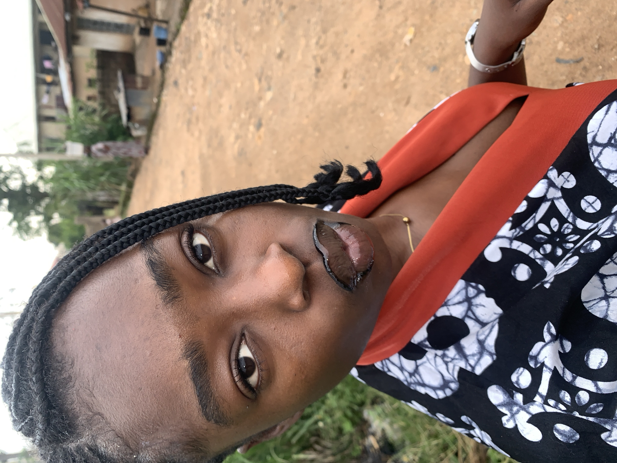 A close-up selfie shows a young Black woman in the foreground, her face turned towards the camera with a pouty expression and black lipstick applied. She has braided hair and is wearing a black and white patterned dress with an orange collar. Her left arm, adorned with a silver bracelet, is extended toward the right side of the frame.
In the background, a dirt road stretches out, lined with simple buildings and lush green vegetation. The scene appears to be in a rural or semi-urban setting, possibly a village like Rafin Par, Nigeria, given the presence of traditional housing structures and the dusty, unpaved road. A person can be seen in the distance on the road, carrying a bucket on their head, suggesting daily life activities. The overall atmosphere is casual and candid, capturing a moment in time within the depicted environment.
