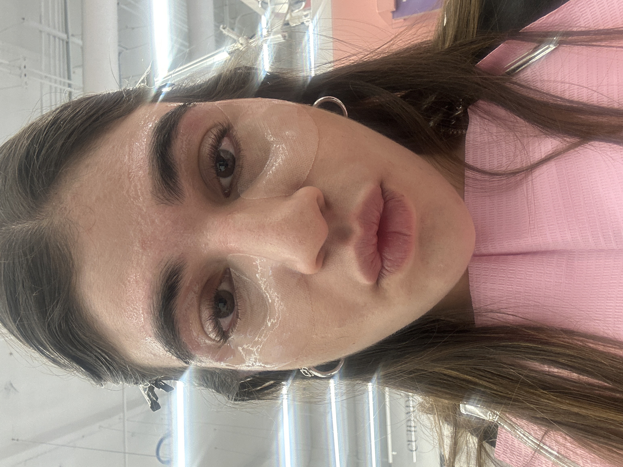 The image captures a female subject from a low-angle, close-up perspective, looking directly at the camera. She is undergoing a cosmetic treatment. Clear gel patches are applied under both eyes, and a translucent substance covers her forehead and cheeks. Long, dark brown hair is secured with silver-toned clips on both sides of her head, pulling it back from her face. Silver hoop earrings are present on both ears. Her lips are slightly parted. A pink, textured disposable drape covers her chest and neck. The background reveals an indoor commercial setting, likely a beauty salon or spa, characterized by white panel ceiling tiles and bright overhead fluorescent lighting. A partially obscured sign in the upper left background displays the text "CUIDADO". This scene is located within the city of Puebla, Mexico.