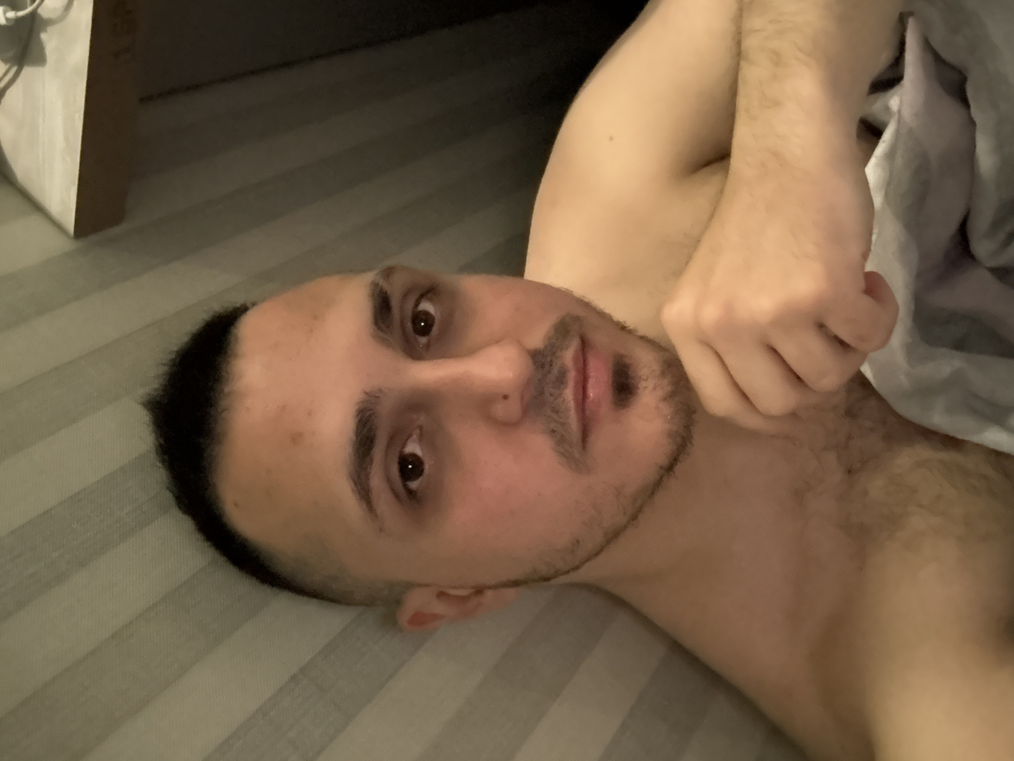 A medium close-up shot shows a male-presenting individual lying down, looking directly at the camera. The person is shirtless, with visible hair on their chest and arms. They have short dark hair and facial stubble or a short beard. Their eyes are dark and wide open, with a direct, somewhat neutral expression. One arm is bent, with the hand resting near the shoulder and partially covered by a light-colored sheet or fabric on the right side of the frame. The individual is positioned on a surface with light-colored, subtly striped sheets, likely a bed or mattress. In the upper left background, there's a glimpse of a light-colored, possibly wooden, piece of furniture or structure. The lighting is somewhat dim, suggesting an indoor setting, possibly a bedroom, during the evening or night. There is no visible text in the image.