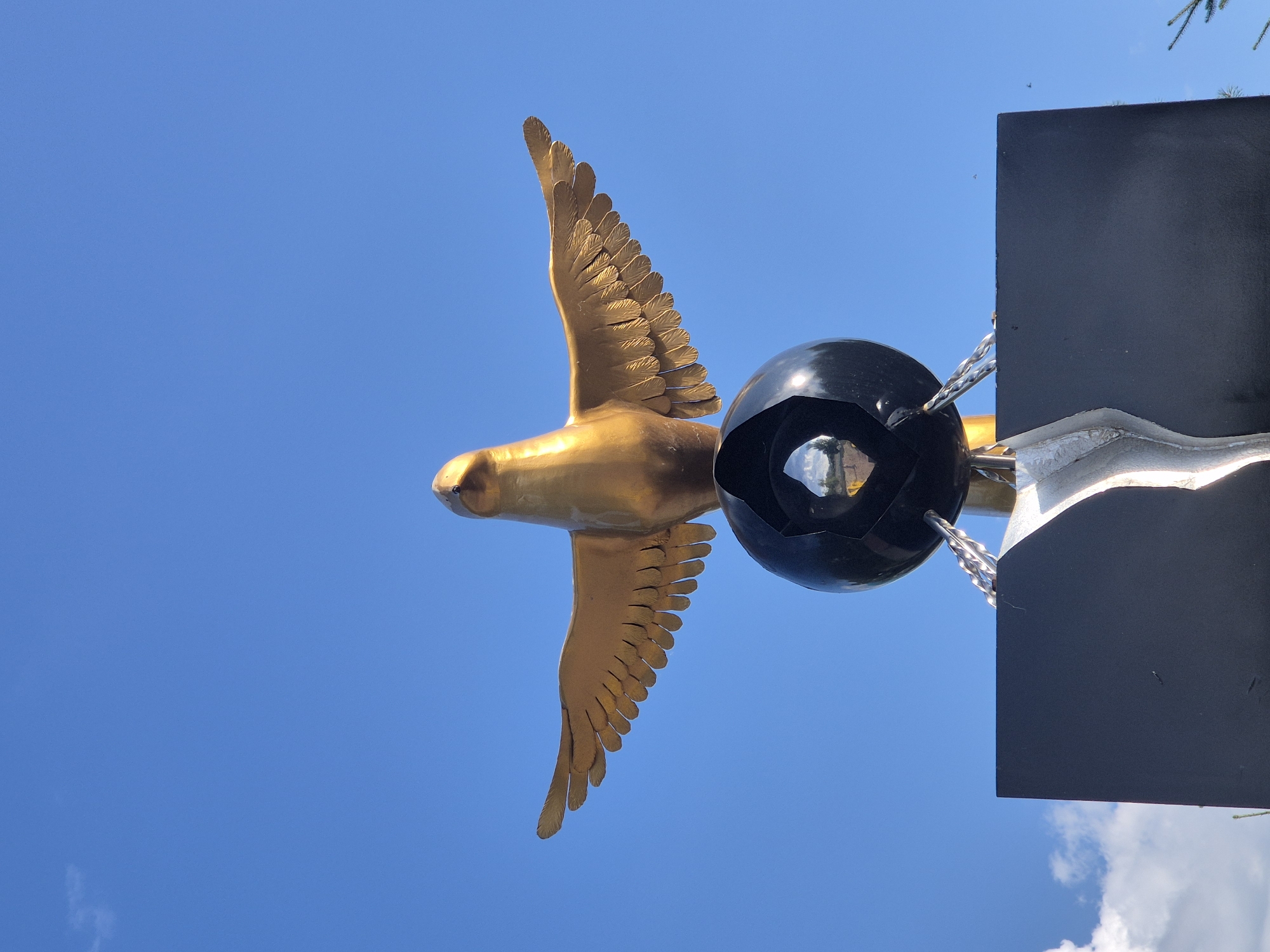 The image shows a close-up, low-angle shot of a golden bird sculpture with its wings spread wide, perched on top of a cracked black sphere. The sculpture is positioned against a clear blue sky with a few wispy clouds visible. The bird has a sleek, modern design, and its golden finish reflects the sunlight. The black sphere appears to be a fountain, as water is seen streaming from its opening and flowing down the sides. Below the sphere is a black rectangular base with a silver, abstract metallic element attached to its front. The sculpture appears to be outdoors, possibly in a public park or square. This sculpture is known as the "Golden Bird" monument and is located in Kokshetau, Kazakhstan.