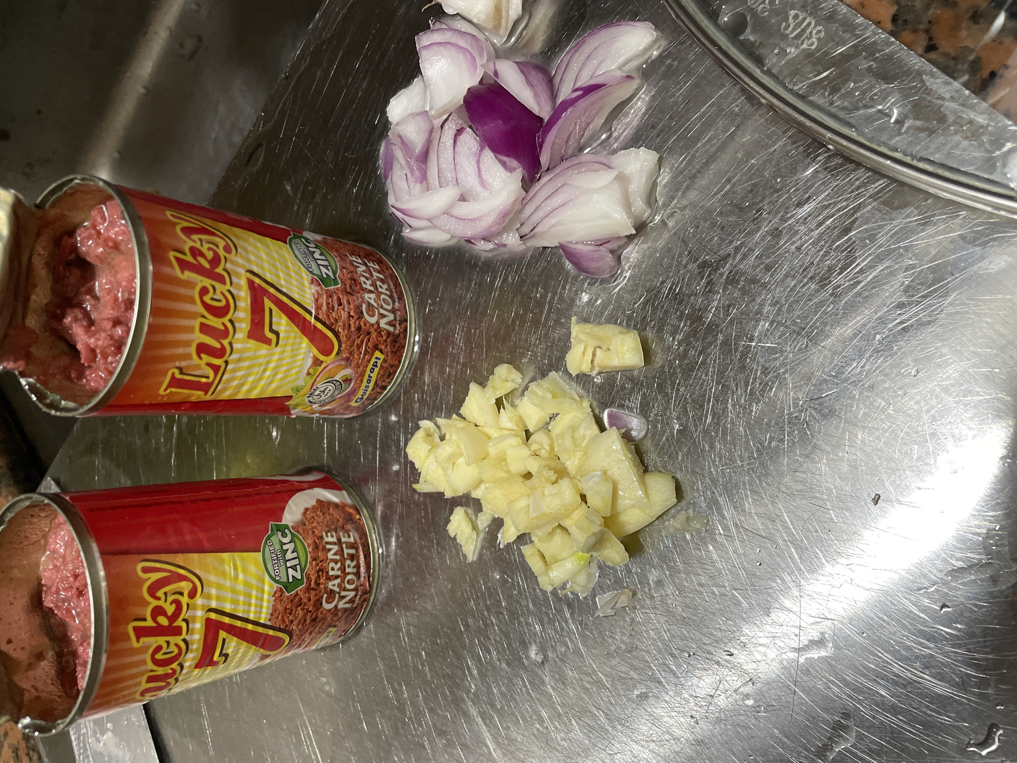 The scene depicts a kitchen preparation area, identified as being in Davao City, Philippines. The primary surface is a highly reflective, scratched stainless steel countertop exhibiting numerous water droplets and streaks. Two open, red and yellow cans of "Lucky 7 Carne Norte" corned beef, each labeled "FORTIFIED WITH ZINC," are prominently positioned towards the top of the frame, revealing the reddish, shredded contents within. To the left of the cans, a pile of coarsely chopped garlic cloves is arranged on the stainless steel. To the right, a separate pile of roughly chopped red onion segments, displaying white and purple layers, is present. A section of a speckled brown and black granite countertop is visible in the lower right corner, partially obscured by a circular glass lid with a metallic rim, which displays "SUS" text. The background features a dark, indistinct area, possibly indicating a sink or adjacent counter space. No individuals are visible in the image, and the overall context suggests active food preparation.