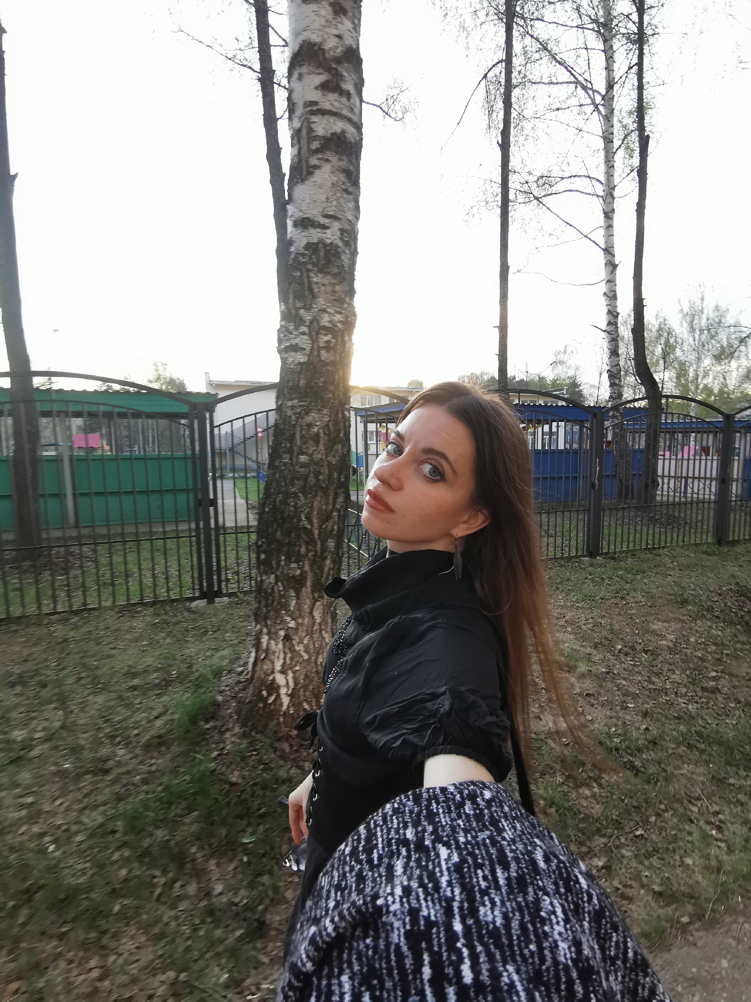 A woman stands outdoors near a tree, likely in a park or residential area within the city of Unknown. She is wearing a black top with puffed sleeves and a patterned sweater, possibly taking a selfie. Behind her, a fence surrounds a building or structure, with additional trees and greenery visible in the background. The lighting suggests it might be early morning or late afternoon. The ground appears to be a mix of grass and dirt.
