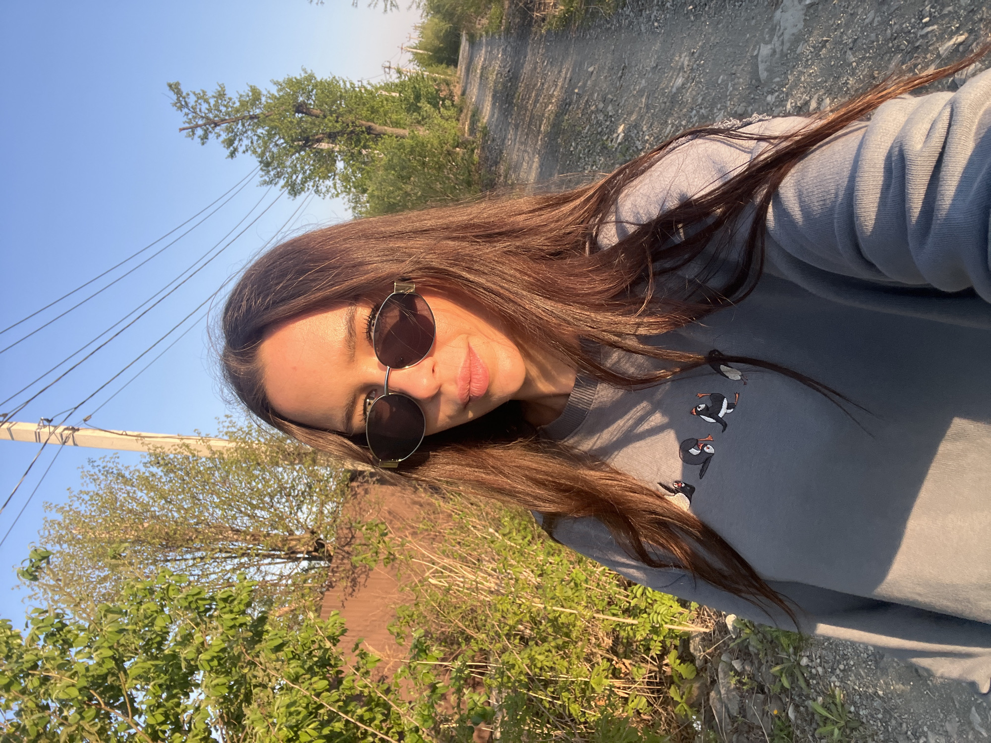 The image depicts a woman wearing sunglasses and a grey sweatshirt with puffins embroidered on it. She appears to be standing on a dirt road in a rural area, with vegetation and trees visible on either side. The sky is blue. Based on the metadata, the location is listed as Unknown, Russia. There are power lines visible in the background.