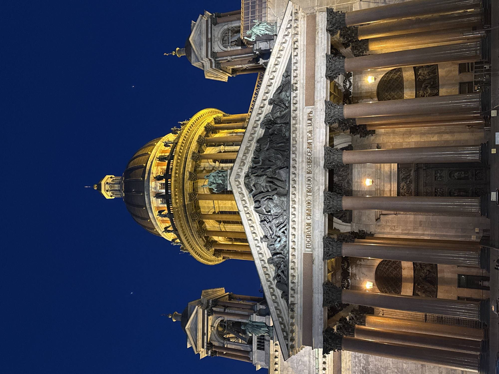 This striking image captures the grandeur of St. Isaac's Cathedral in Saint Petersburg, Russia, at night or late dusk. The massive neoclassical building dominates the frame, illuminated by warm artificial lights that cast a golden glow on its architectural features against a deep, clear blue sky.
The most prominent feature is the large, golden dome, crowned with a smaller cupola and spire, glowing brightly. Below it, a ring of sturdy columns encircles the drum. The main facade features a monumental portico supported by numerous massive columns, likely made of granite or marble. Above these columns, a triangular pediment is filled with intricate sculptural reliefs depicting various figures. Additional statues and ornate architectural details, including elaborate column capitals and decorative friezes, adorn different levels of the building.
The deep blue sky is almost completely dark, with a single bright celestial body, possibly a star or planet, visible in the upper right quadrant. No specific activity is taking place; the scene focuses on the static beauty of the illuminated architecture.
A notable detail is the Cyrillic inscription visible on the frieze below the pediment. The text reads: "ГОСПОДИ СИЛОЮ ТВОЕЮ ВОЗВЕСЕЛИТСЯ ЦАРЬ," which translates to "O Lord, by Thy strength the King shall rejoice." The dramatic lighting enhances the imposing and majestic presence of the cathedral, emphasizing its intricate details and grand scale against the nocturnal backdrop.