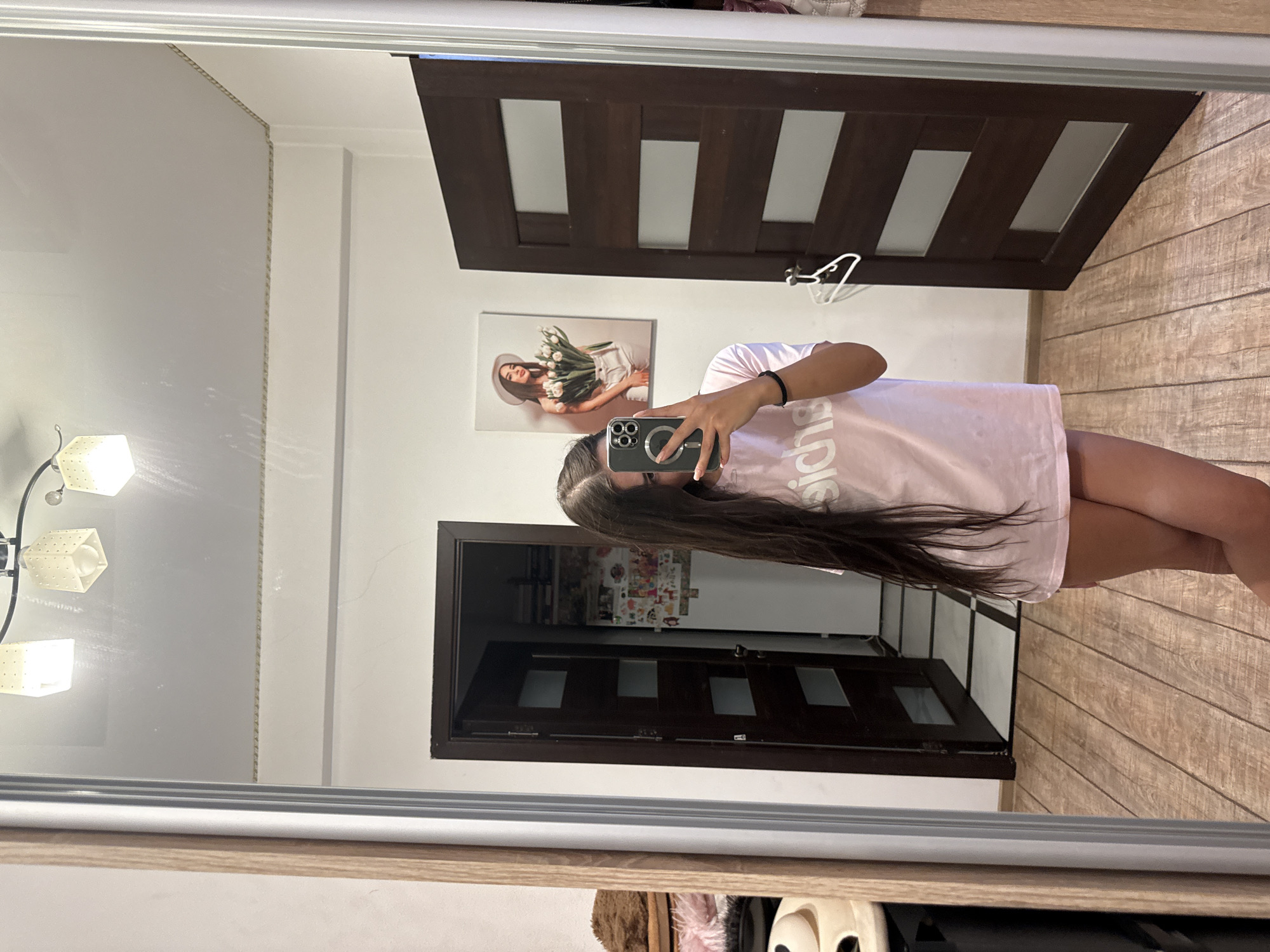 A young woman with long, dark hair is taking a mirror selfie in a room. She is wearing an oversized light pink t-shirt and no visible bottoms, with her legs visible below the t-shirt. She is holding a smartphone in her right hand. The room appears to be a bedroom or dressing area. To her left, a large wooden-framed mirror reflects the scene. Behind her, there are two dark wooden doors with frosted glass panels, one slightly ajar revealing a glimpse of a hallway. On the wall to the right of the open door, a framed picture of a woman wearing a hat and holding tulips is hanging. The floor is covered in light-colored wooden planks. The lighting suggests indoor illumination, possibly from overhead fixtures. The specific location within Chișinău, Moldova cannot be determined from the image.