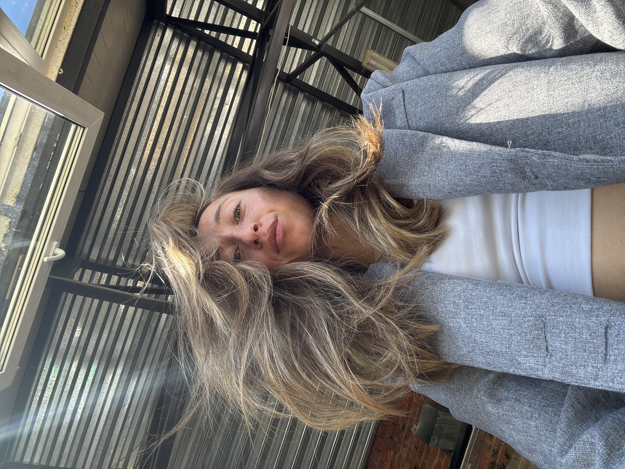 A low-angle, medium shot captures a woman from the chest up, facing the camera outdoors. She has blonde hair, which is being blown across her face by the wind, obscuring part of her features. She is wearing a gray blazer over a white tank top. Her left arm is bent at the elbow and resting against her body, while her right arm is not visible. The background consists of a corrugated metal roof structure with exposed beams, suggesting an urban outdoor setting. The overall lighting indicates it is daytime. The provided image context does not contain information that would place the scene in Chaikovsky, Russia.