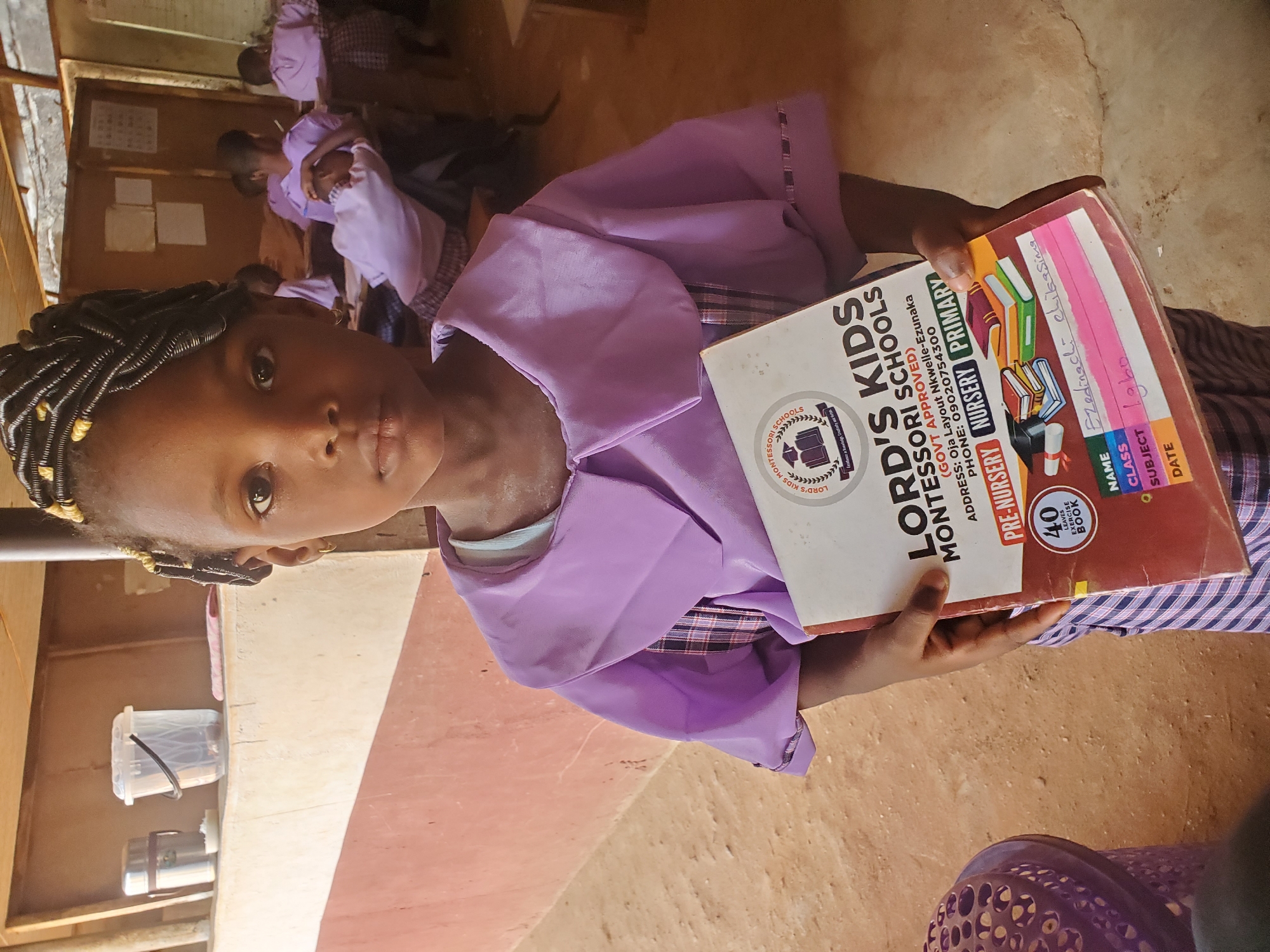 A young girl stands in what appears to be a classroom in Ukwu-Abwa, Nigeria. She is holding a book titled "Lord's Kids Montessori Schools Pre-Nursery Primary". Her name is Ezedinachi Chikasima and her subject is Igbo. She is wearing a purple uniform and her hair is styled in braids. Other children, also in uniforms, are visible in the background. The classroom has a simple, somewhat rustic appearance.