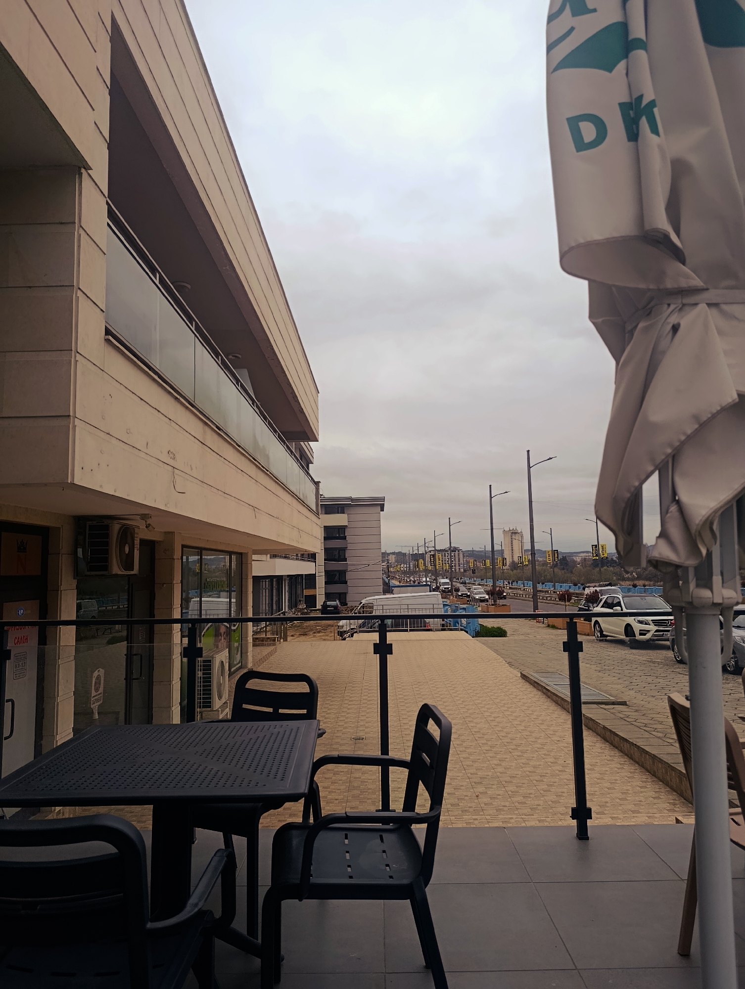 The image depicts an outdoor seating area of a café or restaurant located in Kardzhali, Bulgaria. A black square table with three chairs sits on a gray tiled patio in the foreground. A partially folded umbrella obscures the right side of the frame, revealing the letters "DK" on its fabric. Beyond the patio, a paved walkway runs alongside a multi-lane street with ongoing traffic. Several vehicles, including a white Mercedes-Benz, are visible. Streetlights line the road, and in the distance, buildings are discernible against an overcast sky. The café is part of a multi-story building with a tan facade and glass-paneled balconies. Overall, the scene conveys a quiet, urban setting on a cloudy day.