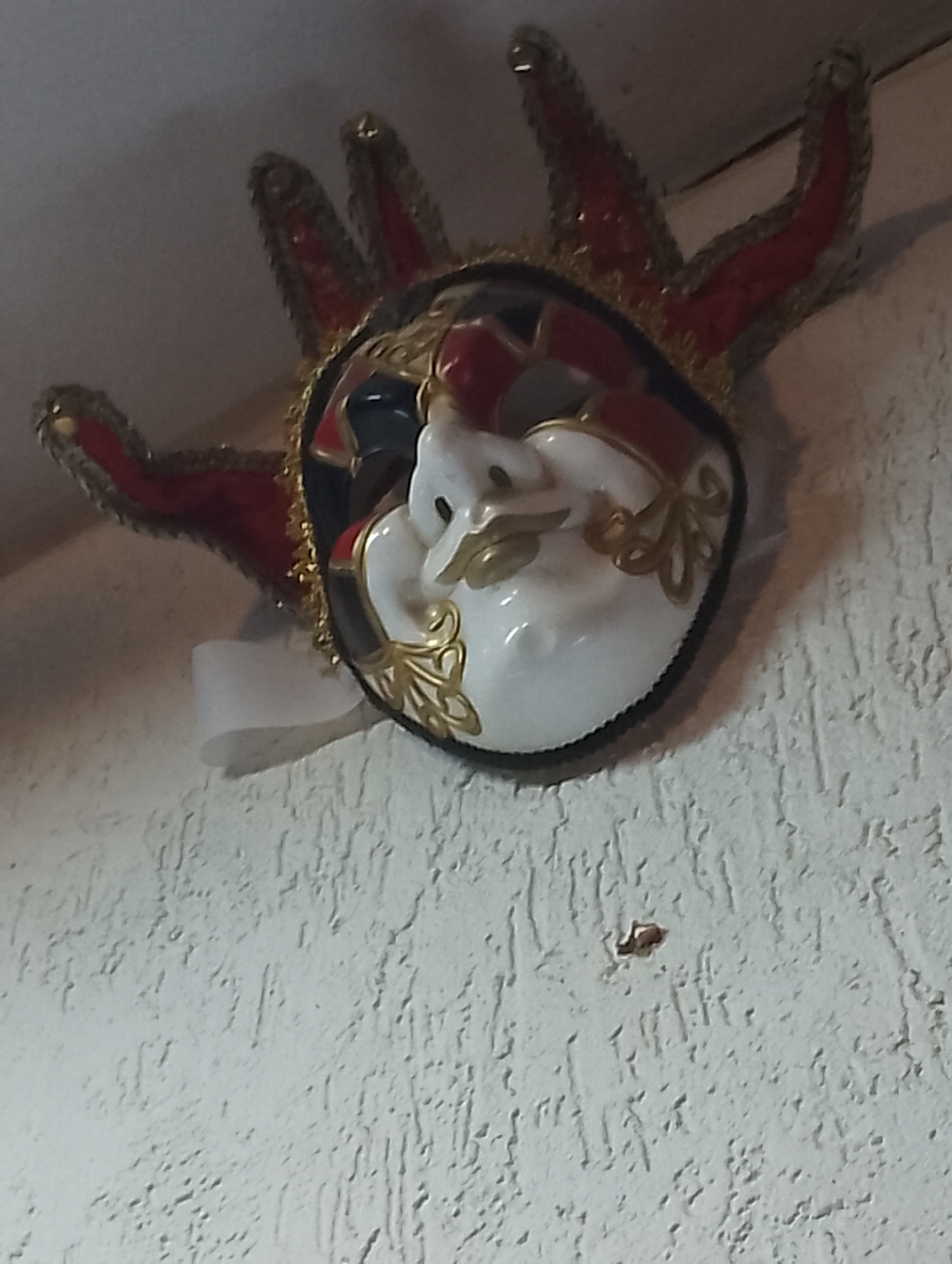 A mask with five red horns, trimmed with gold, is mounted on a textured white wall. The mask's face is white with gold filigree around the eye area and a comical, puckered expression. Red and black accents are painted around the border of the face, inside the horn base. A small piece of white ribbon hangs from the side of the mask. The image appears to be taken indoors. The location is unknown.
