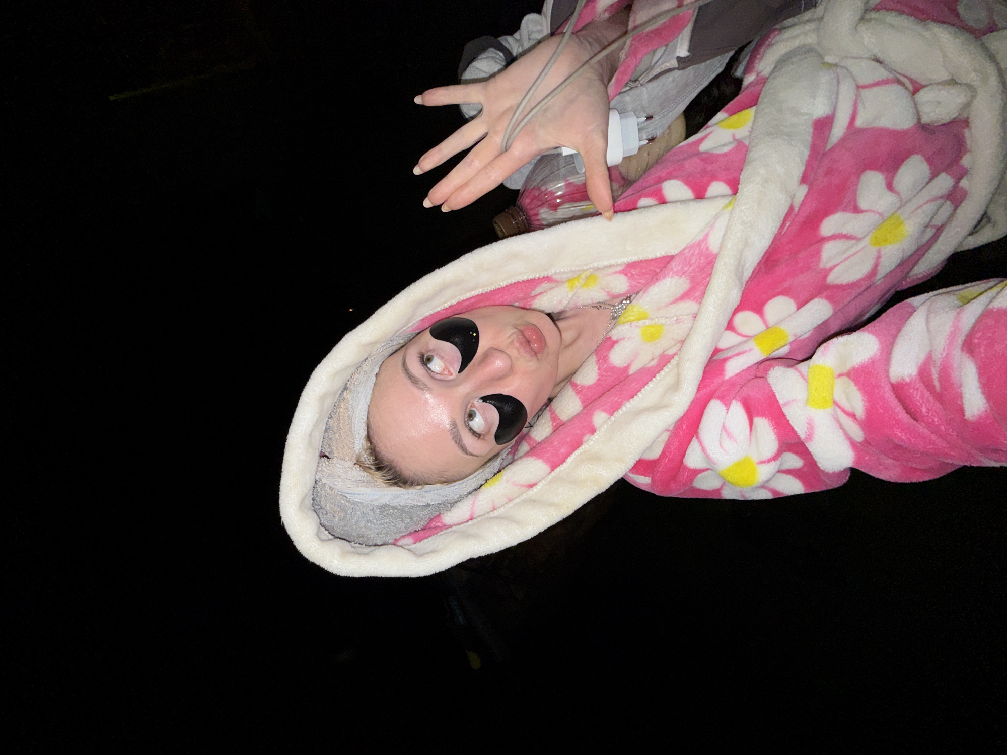 A young woman is pictured from an oblique angle, lying down. She is wearing a pink and white floral patterned robe with a white fluffy lining around the hood. She has a gray towel wrapped around her head and black eye patches under her eyes. Her hand is raised near a phone charger and a plastic bottle. The background is black, suggesting a night setting. The scene is not readily identifiable as being in Nizhnyaya Chermoda, Russia, as there are no specific markers to indicate the location.