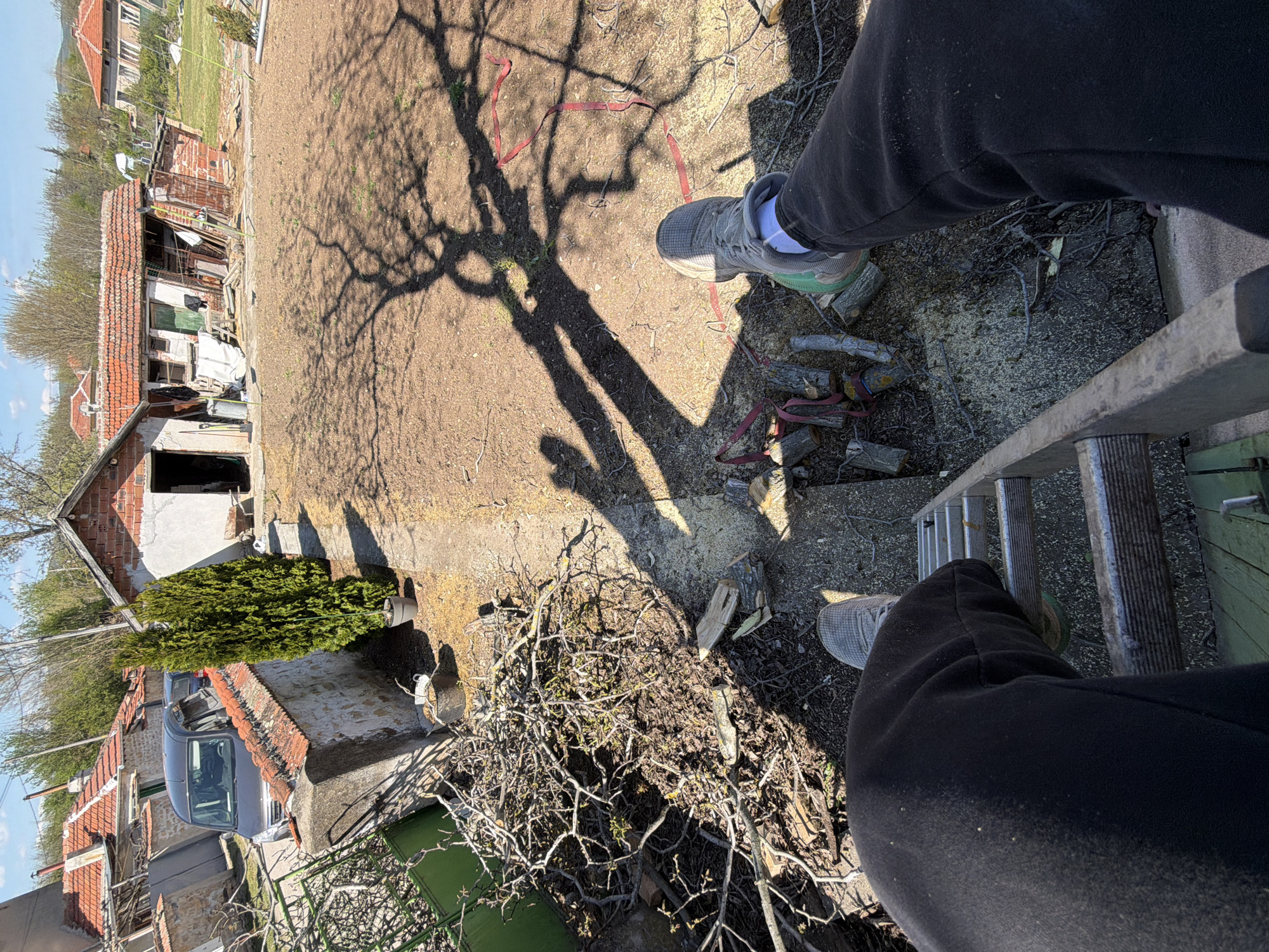 An eye-level shot from a ladder reveals an outdoor scene in Unknown, Unknown, marked by a mix of man-made and natural elements. The lower half of a person wearing dark sweatpants and light-colored running shoes is visible on the ladder. Below the ladder, there are piles of pruned branches. The ground is a mix of dirt and concrete, marked with the long, dark shadow of a bare tree. In the background, there are several buildings with orange tiled roofs, a green evergreen tree, and a parked silver van. The sky is a light blue, suggesting daytime.