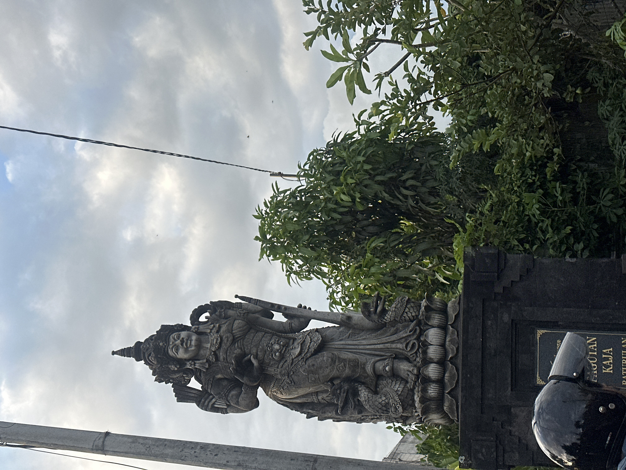 The image shows a stone statue of a deity standing in Sukawati, Indonesia. The statue is intricately carved with ornate details and is surrounded by lush green trees and foliage. The sky is overcast with clouds. A black motorcycle helmet is partially visible on the right side of the frame, suggesting a person is present but not fully visible. A sign next to the statue has the words "PAGULAN KAJA BATUBULAN." A powerline is also visible against the cloudy sky.