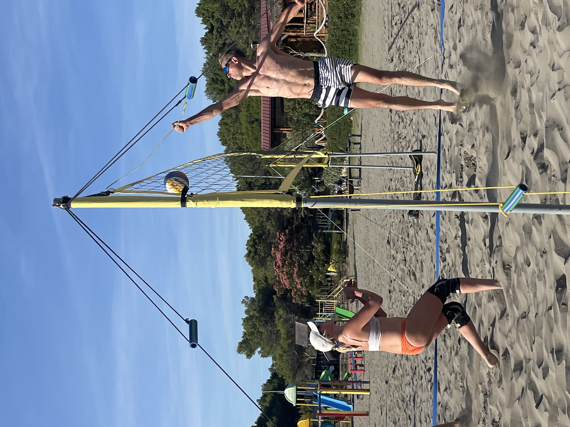 This is a full, eye-level, outdoor, natural light, horizontal shot of two people playing beach volleyball. The scene takes place on a sandy beach with a clear blue sky above and trees in the background. On the left, a woman wearing a white bikini top, orange bottoms, and knee pads is in a defensive stance, looking up at the ball. On the right, a man in black and white striped shorts is mid-air, spiking the volleyball over the net. The volleyball net is yellow with a black net. There are playground structures visible in the background, suggesting a recreational area. The overall environment appears to be a sunny day at the beach, likely in a resort or park area. The image captures a dynamic moment of athletic activity.