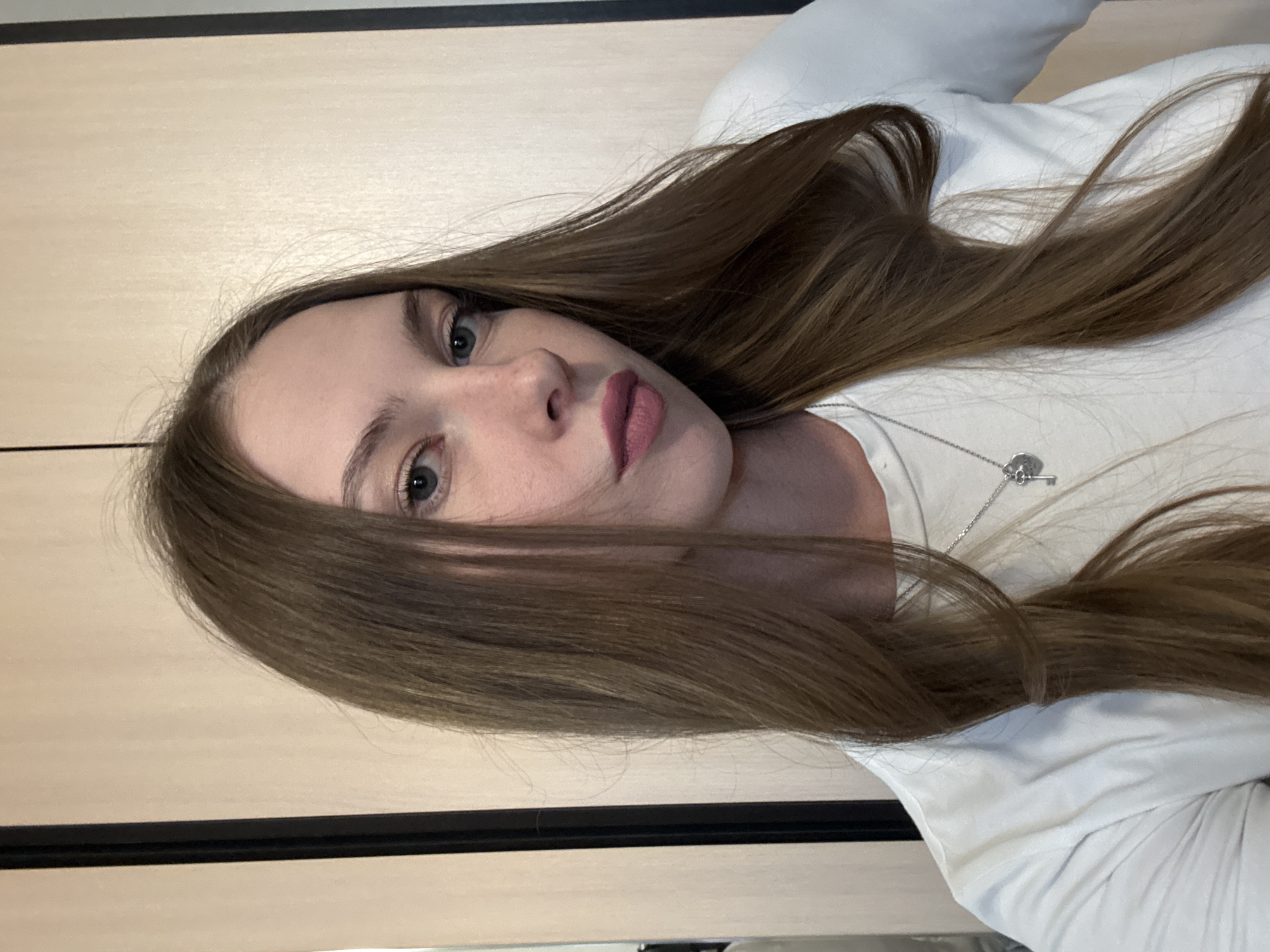 The media file is a portrait of a young woman with long, light brown hair. She is wearing a white long-sleeved shirt and a silver necklace with a key pendant. Her makeup includes lipstick in a muted pink shade. She is looking towards the camera with a neutral expression. The background appears to be a room with light-colored wooden paneling and a dark horizontal line, possibly a baseboard or the edge of a piece of furniture. The lighting is even, suggesting an indoor setting, and there are no strong shadows. The image is oriented vertically. Based on the provided location context of Perm, Russia, the setting is likely an indoor environment within that region. There is no visible text in the image.