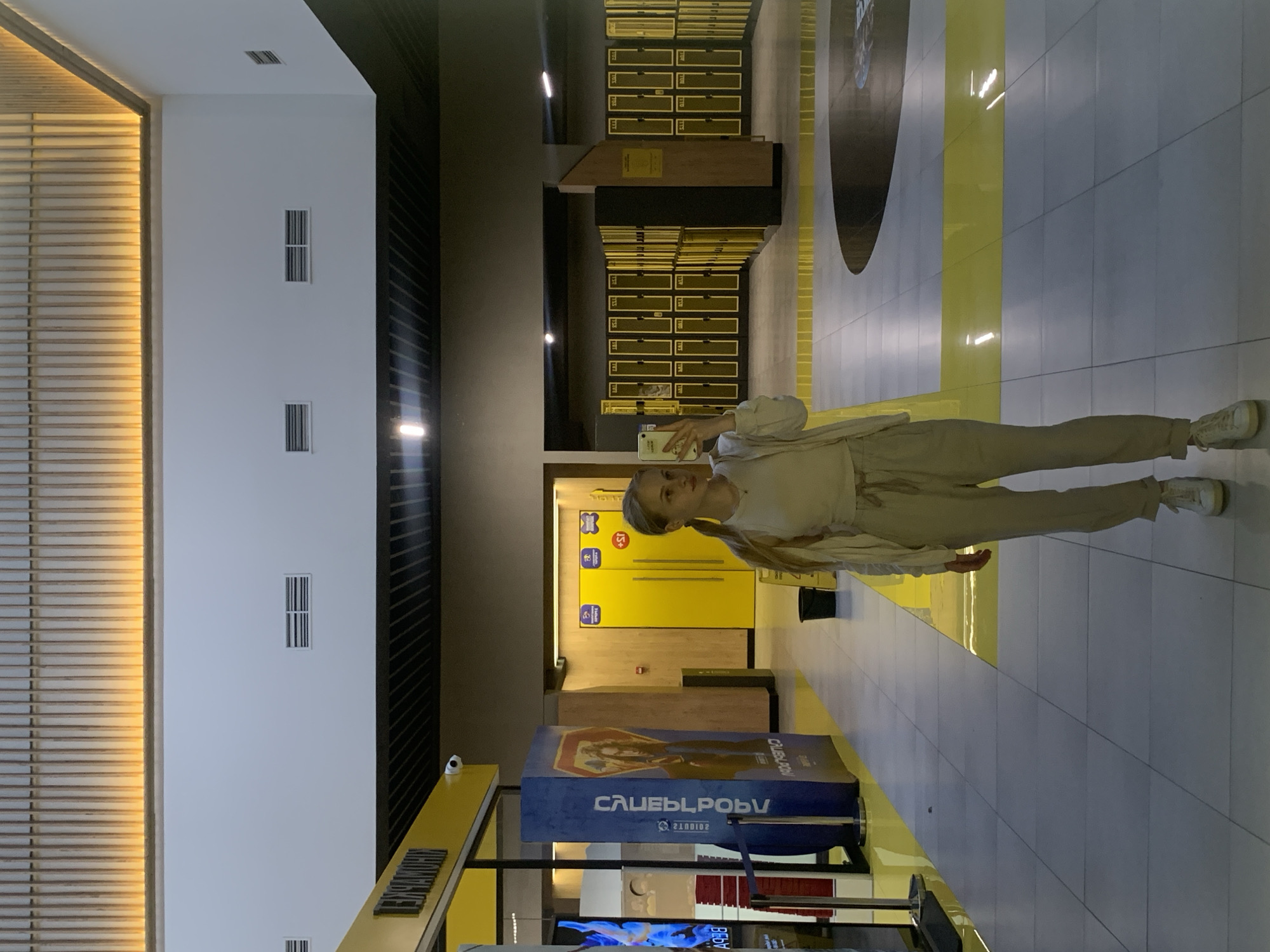 The image depicts a young woman taking a selfie in a modern, brightly lit indoor public facility located in Kyiv, Ukraine. The woman stands centrally in the frame, holding a white smartphone with her right hand to capture her reflection. She has light-colored hair tied in a ponytail and is dressed in a light-colored (beige/cream) outfit consisting of a t-shirt, an open cardigan or jacket, loose-fitting trousers, and white sneakers. Her gaze is directed at the smartphone screen.
The facility features a contemporary design with light gray tiled floors intersected by wide yellow stripes. A large circular dark gray design is visible on the floor to the right. The ceiling integrates warm yellow-lit wooden slats along its edges with plain white and dark gray suspended sections, which contain rectangular air vents and strip lighting.
To the left of the woman, a prominent yellow structure displays the Cyrillic text "ЛЕКСНАМОЯН" in black capital letters. Adjacent to this is a blue pillar featuring mirrored Cyrillic text and what appears to be a digital display screen showing blue and white graphics. Red stanchions with ropes delineate a queuing area in this section. A small black security camera is mounted on the yellow structure.
To the right, a long wall is lined with numerous bright yellow lockers, suggesting a changing room or storage area. Further back, two yellow doors are visible, marked with symbols resembling gender signs and "+1S" or "+18" icons. Wooden paneling covers sections of the walls in this locker area. A black bucket rests on the floor near the lockers. The polished floor reflects the overhead lighting and yellow elements of the environment.