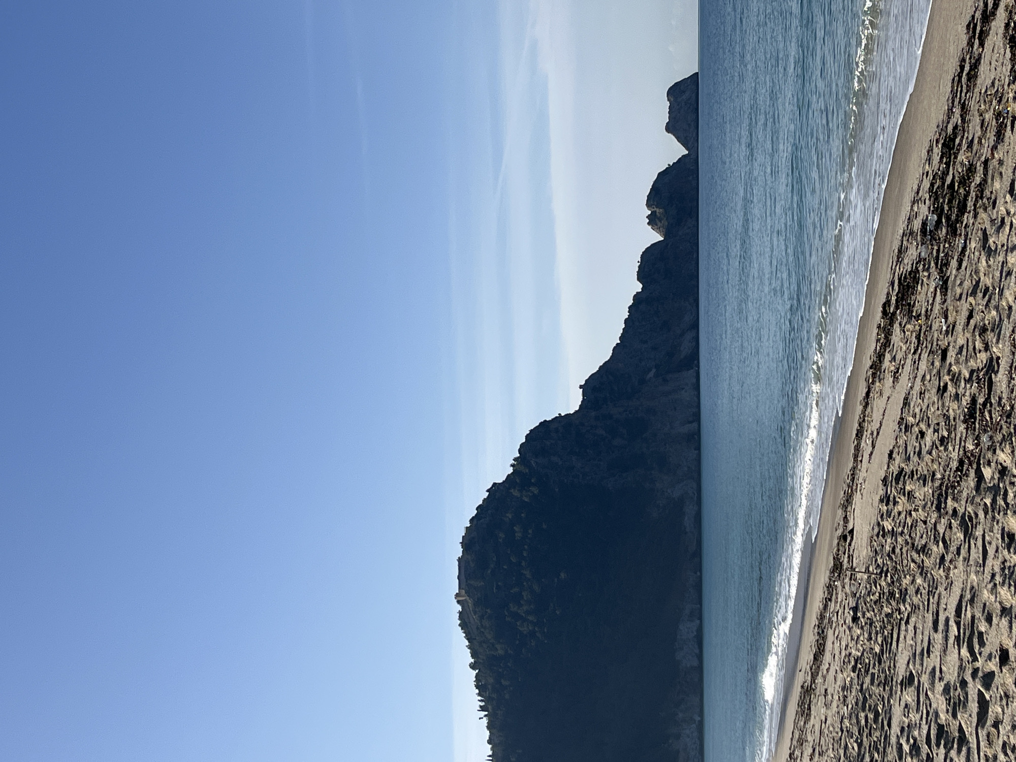 A landscape photograph depicts a coastal scene in Alanya, Turkey, featuring a beach, the Mediterranean Sea, and a mountainous headland. The image is oriented vertically. The left half of the image is dominated by a clear, blue sky with faint wisps of clouds. In the middle ground, a large, dark-colored headland rises from the sea. The right side of the image shows the beach, comprised of sand and organic debris. Waves from the sea gently lap the shore. The overall scene suggests a tranquil, natural setting.