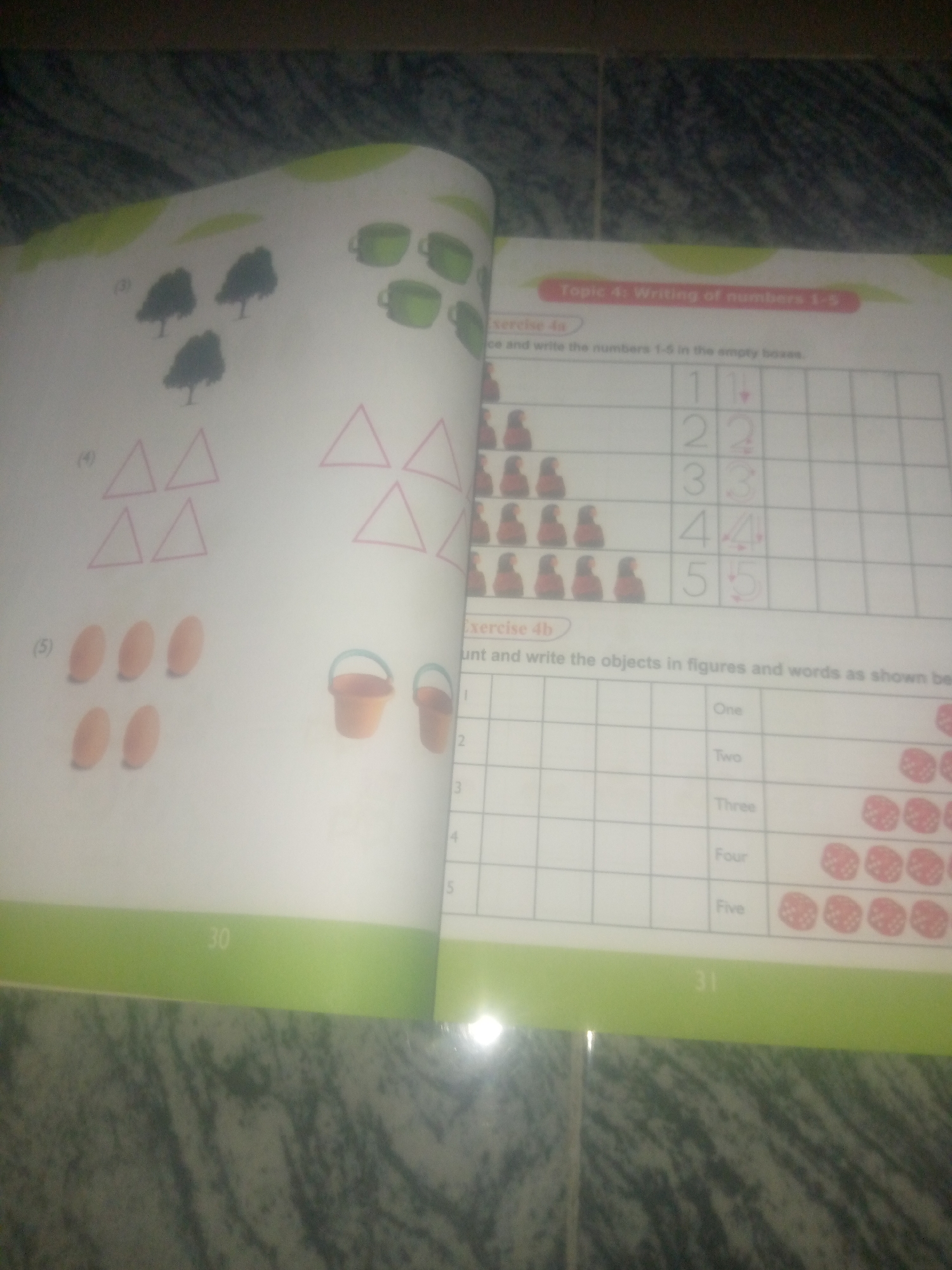 The image displays an open workbook with pages on the right and left. The right page features "Topic 4: Writing of numbers 1-5" and "Exercise 4a" with instructions to trace and write numbers 1-5 in empty boxes, accompanied by rows of figures. Below this is "Exercise 4b" with instructions to count and write objects in figures and words, showing rows of numbers, words (One, Two, Three, Four, Five), and images of dice. The left page shows illustrations of trees, triangles, eggs, and buckets, with numbers (3), (4), and (5) next to them. The workbook pages are laid flat on a dark, textured surface, possibly a table, with the bottom edges of the pages showing page numbers "30" on the left and "31" on the right. The image is a close-up, top-down shot, with no elements suggesting a specific location within the city of Jauro Yinu, Nigeria.