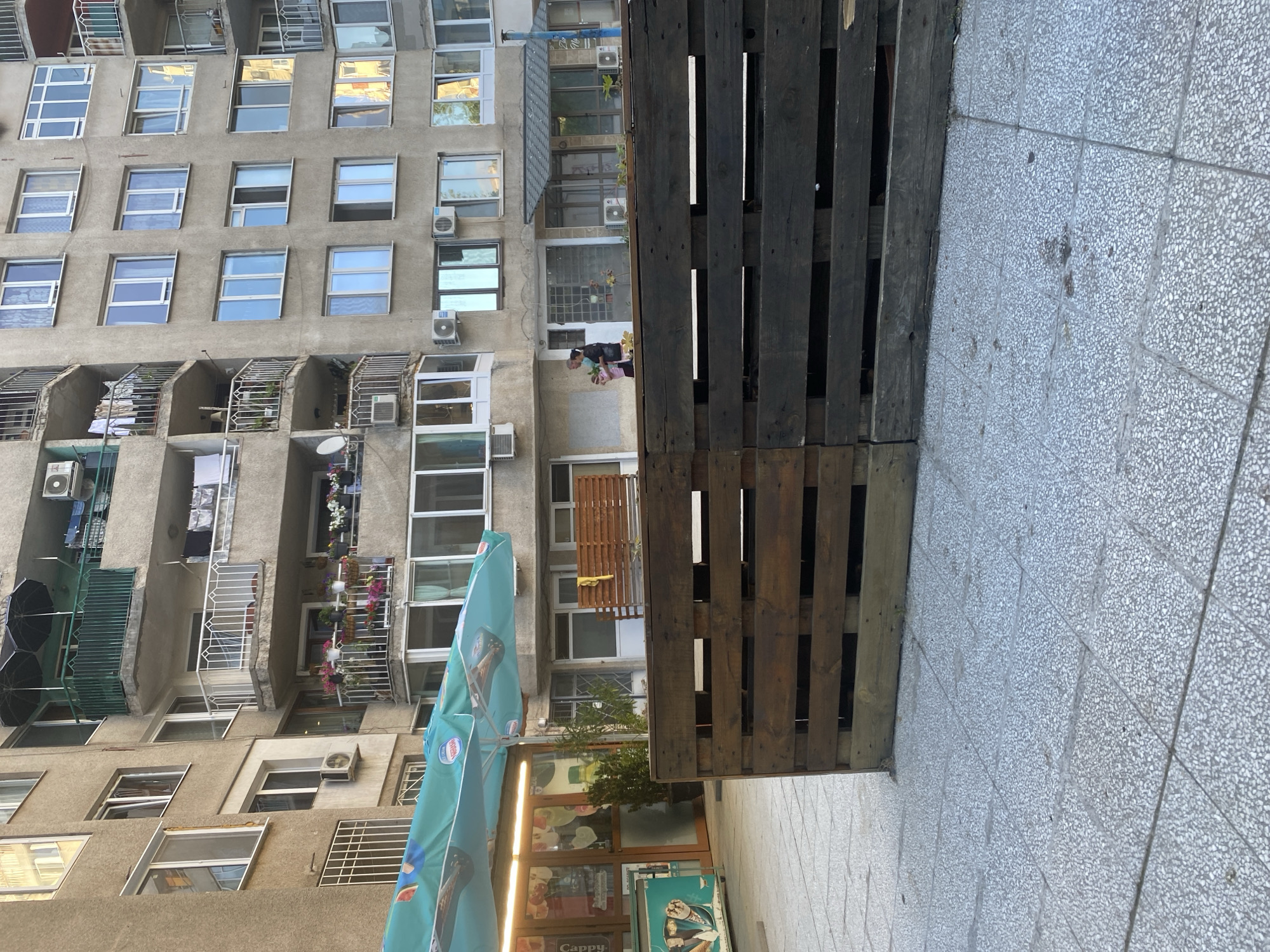 The scene is an urban environment in Sofia, Bulgaria. A tall, Soviet-era apartment building dominates the left side of the image. The facade is gray, with numerous windows, balconies of different designs, and visible air conditioning units. Laundry is hanging on some balconies.
On the right is a sidewalk paved with small, speckled gray tiles. A structure made of dark wooden pallets is positioned between the building and the sidewalk.
In the lower portion of the frame, there is a storefront with advertisements, and a blue parasol.
A person is visible on one of the balconies, wearing a pink outfit.