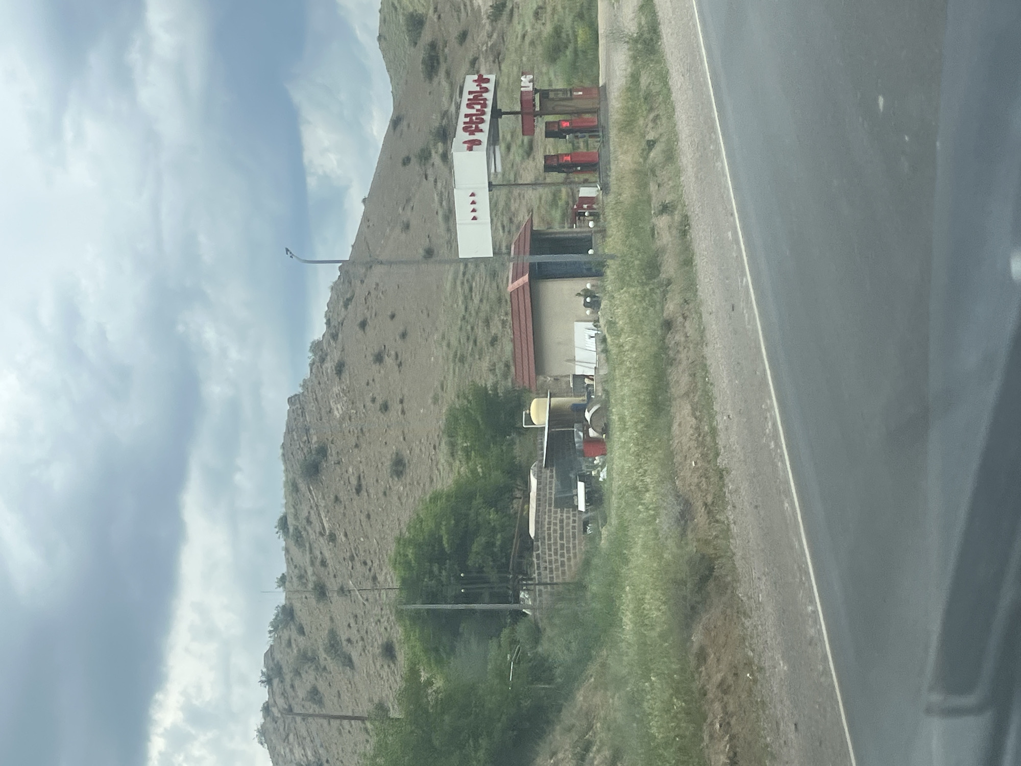The image displays a roadside scene featuring a gas station or similar establishment set against a mountainous, arid backdrop under a cloudy sky. The photograph is oriented vertically, with the road running along the right side and the large hill occupying the left and background.
On the right, a paved road stretches, bordered by a narrow strip of grass and dry earth. To the left of the road, a significant portion of the frame is taken up by a large, rocky hill or mountain, covered with sparse, dry vegetation.
In the mid-ground, a prominent white sign with red letters stands. The visible Armenian text on the main sign reads "ՆԱՎԹԱՅԻՆ ԲԱԶԱ," which translates to "Oil Base" or "Petroleum Base," indicating a fuel station. Below this, another section of the sign displays a series of red diamond or triangular shapes. Several red fuel pumps are visible directly beneath the main sign structure. Adjacent to the pumps, there's a building with a reddish-brown roof, possibly an office or convenience store, and an attached covered area. Further to the left, nestled among some green trees and bushes, is another smaller structure with a brick base, along with various pieces of equipment, including a distinctive yellow cylindrical object.
The sky is overcast with multiple layers of clouds, suggesting a cloudy day. The overall setting appears to be a functional, utilitarian stop in a relatively remote and rugged, hilly region, consistent with the location context of Chiva, Armenia. The photo seems to have been taken from a moving vehicle.