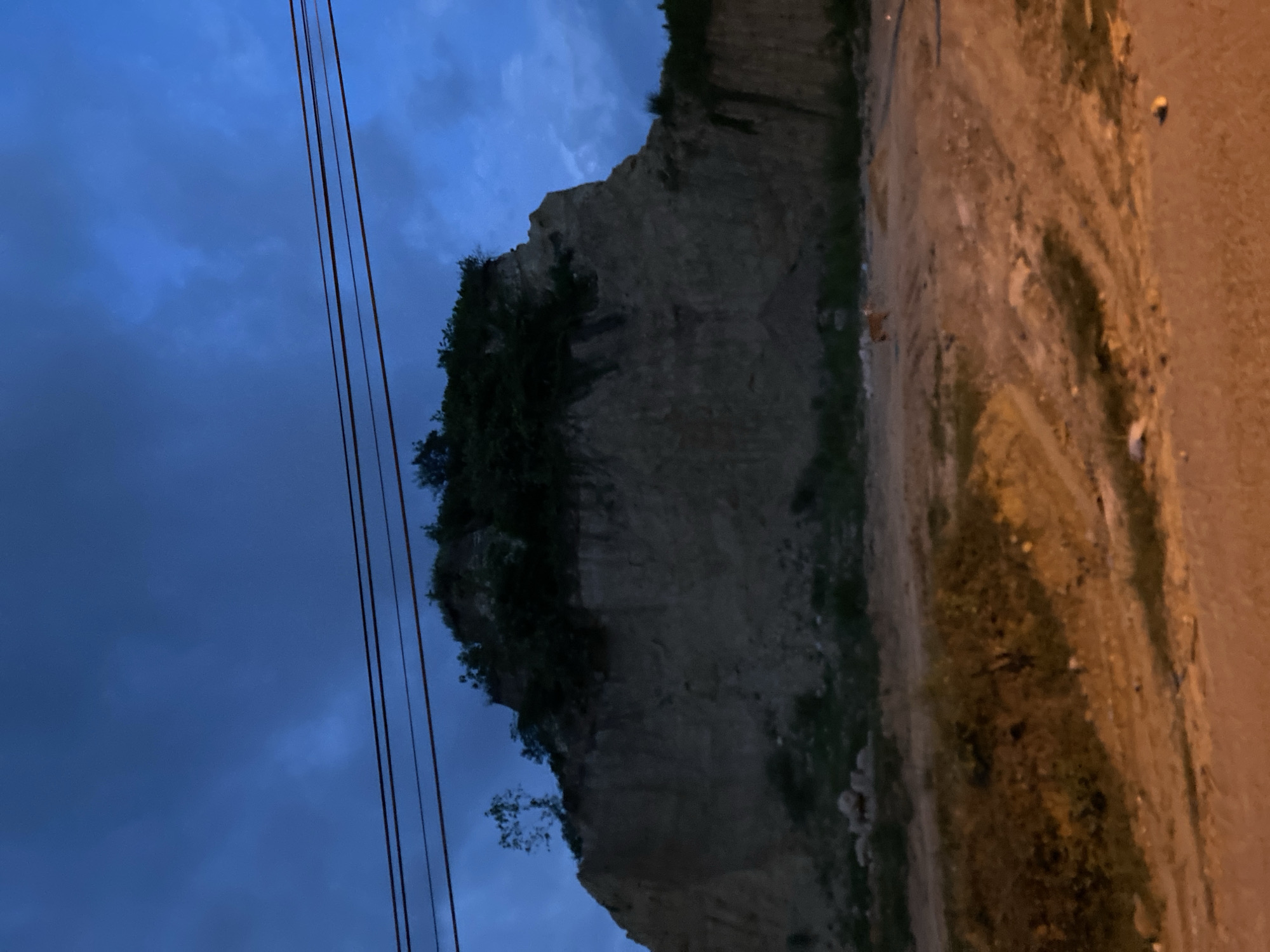 The image shows a cliff face adjacent to a dirt road in Lhokseumawe, Indonesia. The sky above is a dark blue with clouds. Power lines are visible in the upper left. Vegetation grows on top of the cliff and partially down the face. The dirt road is on the right side of the frame. The image appears to have been taken at dusk or dawn.