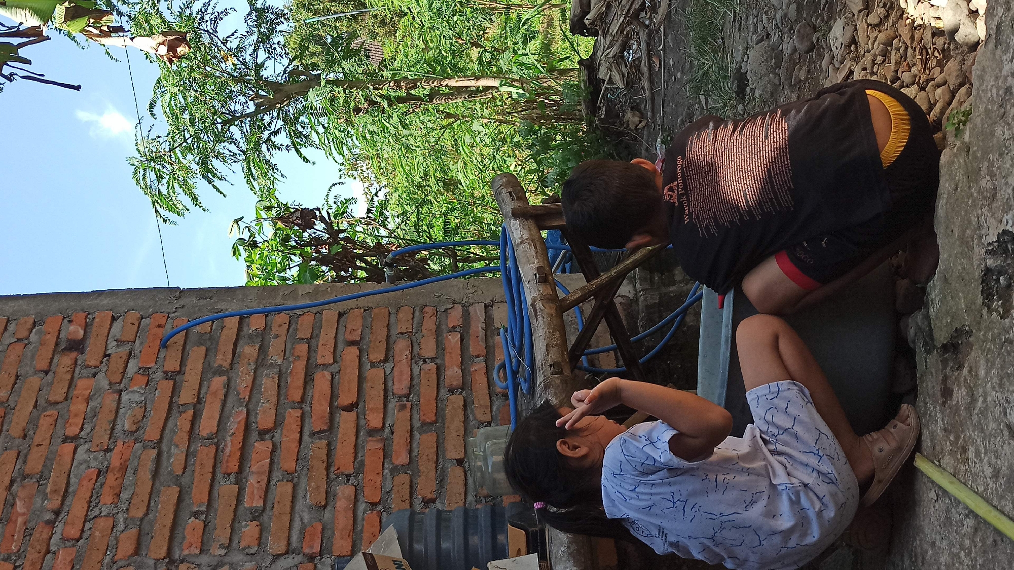 In Gegeran, Indonesia, two children, a girl with a ponytail and a boy, squat in front of a large gray basin outdoors. The girl, facing away from the camera and to the left, wears a white t-shirt with a blue pattern and white shorts, with one hand near her face. The boy, facing away from the camera and to the right, wears a black t-shirt with a dense block of text in a lighter color on the back and black shorts with red and yellow trim. They are positioned on a rough, rocky ground. Behind them, a brick wall with visible mortar rises vertically. A blue hose snakes across a wooden makeshift table structure, also visible behind the children, and appears to be connected to something on the wall. Green trees and foliage dominate the background under a bright blue sky with a few wispy clouds. Various objects, including a large water dispenser and a clear plastic container, are present near the brick wall. The scene appears to be in a residential outdoor area.
