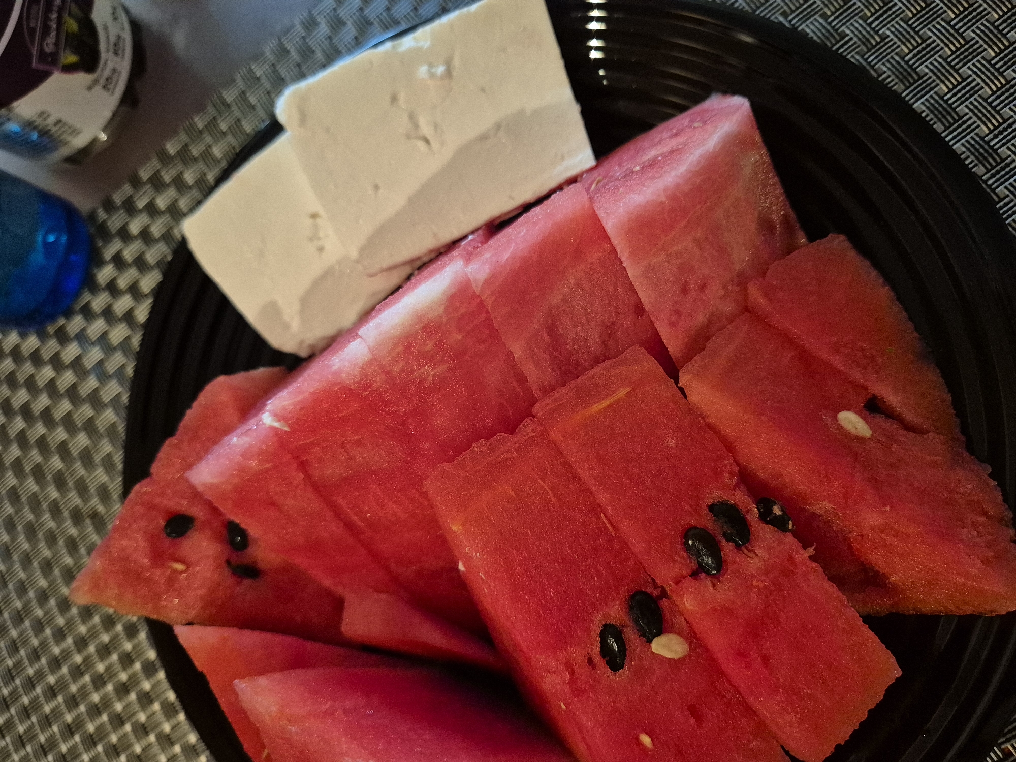 A top-down shot captures a serving of sliced watermelon and a block of white cheese on a black plate, which rests on a gray and black woven placemat. The watermelon, cut into triangular and rectangular wedges, displays vibrant red flesh with scattered black seeds and a few pale, underdeveloped seeds. The white cheese, possibly feta, is presented as a solid, irregular block, contrasting sharply with the red fruit. The black plate has subtle concentric ridges. In the upper left background, part of a dark blue glass or bottle is visible, alongside a jar with a purple label that partially shows the word "Blackberry" and some blurred text below it. The setting appears to be indoors, likely a table, suggesting a casual snack or meal.
