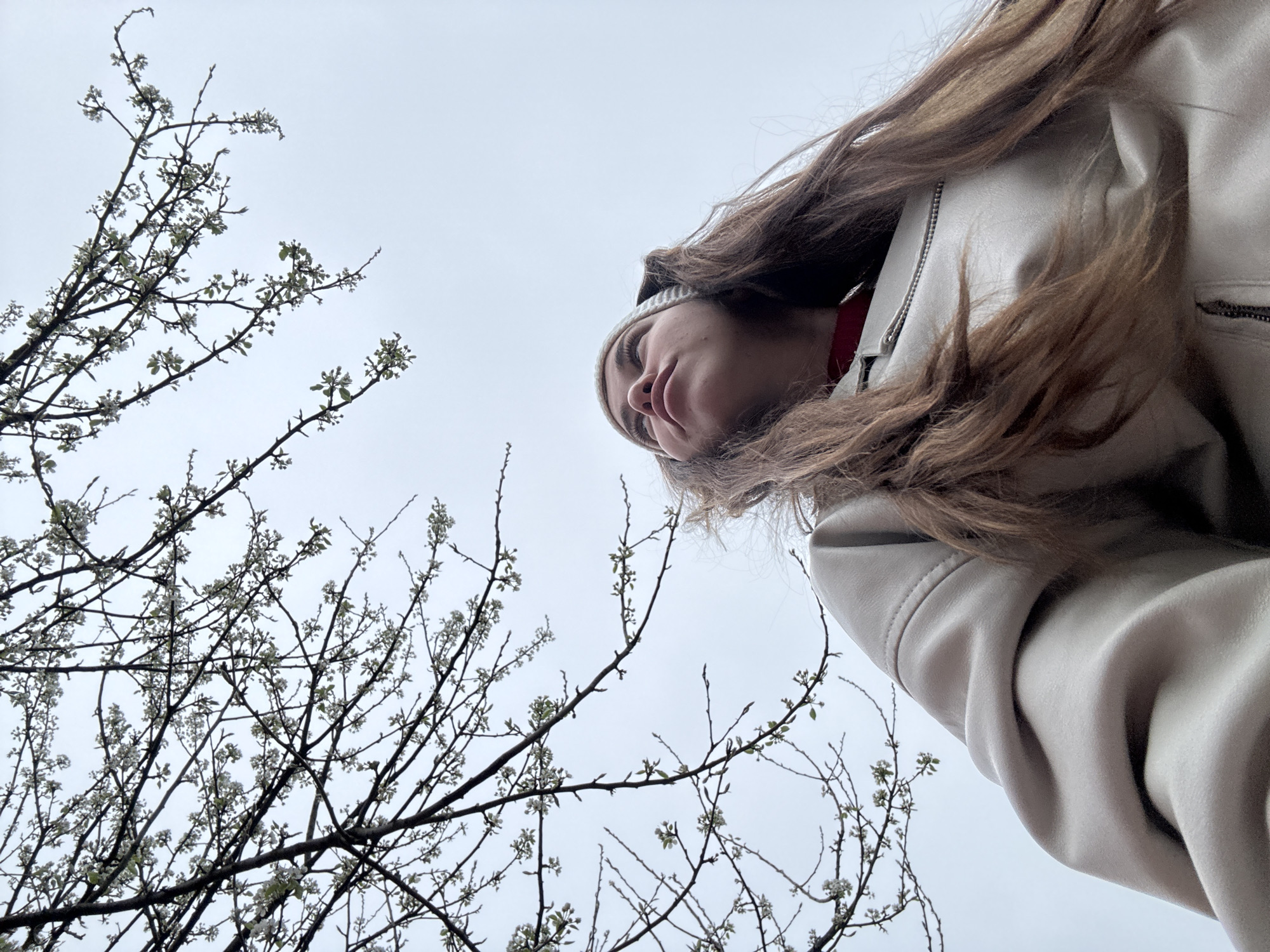 A low-angle shot depicts a person with long, brown hair wearing a white beanie and a light-colored jacket. The person's face is visible, looking upward. In the foreground, branches of a tree with small white flowers are visible against an overcast sky. There is no specific information present to confirm the location as Мещёры, Russia. The scene focuses on the individual's perspective looking up at the tree and sky.