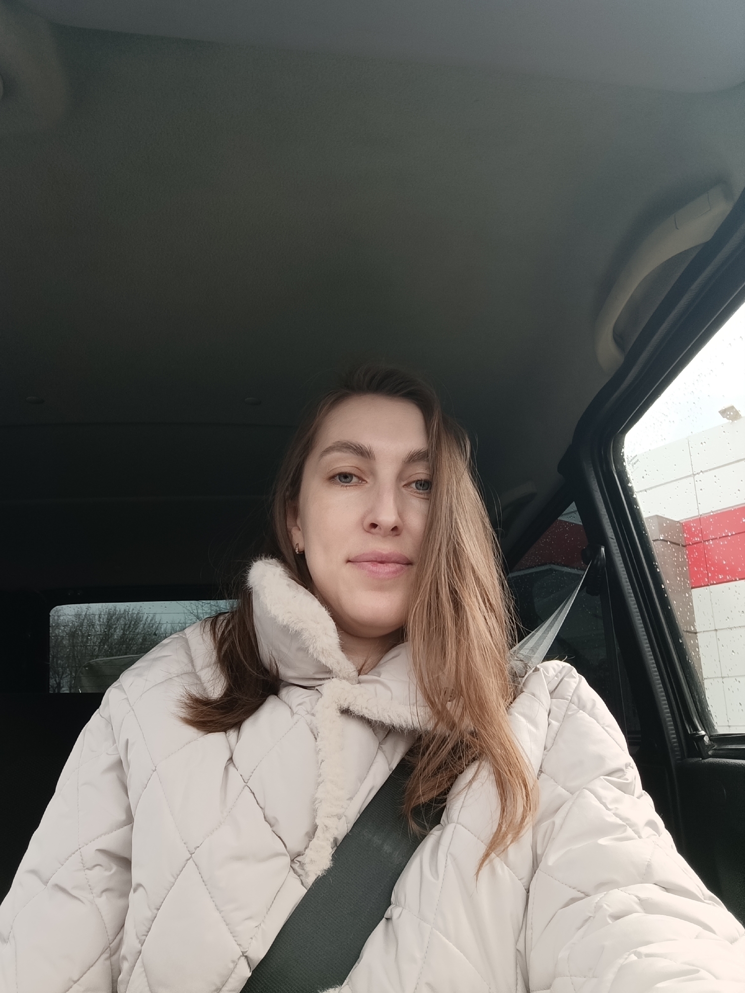 A woman with fair skin, light brown hair, and blue eyes is seated in a car, wearing a light beige quilted jacket with a fuzzy collar and a seatbelt. The car door is open. The background shows a building with white and red panels, and rain is visible on the window. The location is Chaikovsky, Russia.