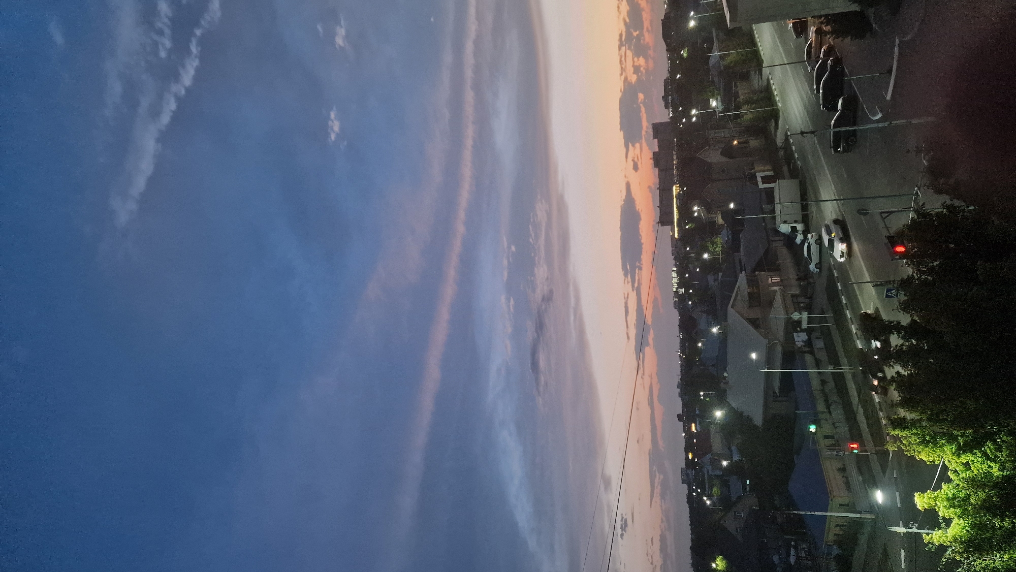 The image depicts a dusk scene in the city of Kostanay, Kazakhstan. The sky is a gradient of deep blue at the top, transitioning to purple and then to a soft orange and pink near the horizon. Wispy clouds are scattered across the sky, some catching the fading sunlight.
Below the sky, a residential area is visible. Houses with pitched roofs, some with visible chimneys and windows, are clustered together. Streetlights are illuminated, casting pools of light on the roads and sidewalks. A road runs through the scene, with several vehicles present. Two cars are actively driving, one white SUV and a darker sedan. Other cars are parked along the side of the road. A truck is also visible. Traffic lights are illuminated, with a red light showing at an intersection. Pedestrian crossing markings are visible on the road.
In the foreground, lush green trees are present, their leaves illuminated by the artificial lights. Overhead power lines are also visible stretching across the frame. In the distance, larger buildings with lights are visible, suggesting a more commercial or denser part of the city. The overall impression is a quiet urban evening in Kostanay.
