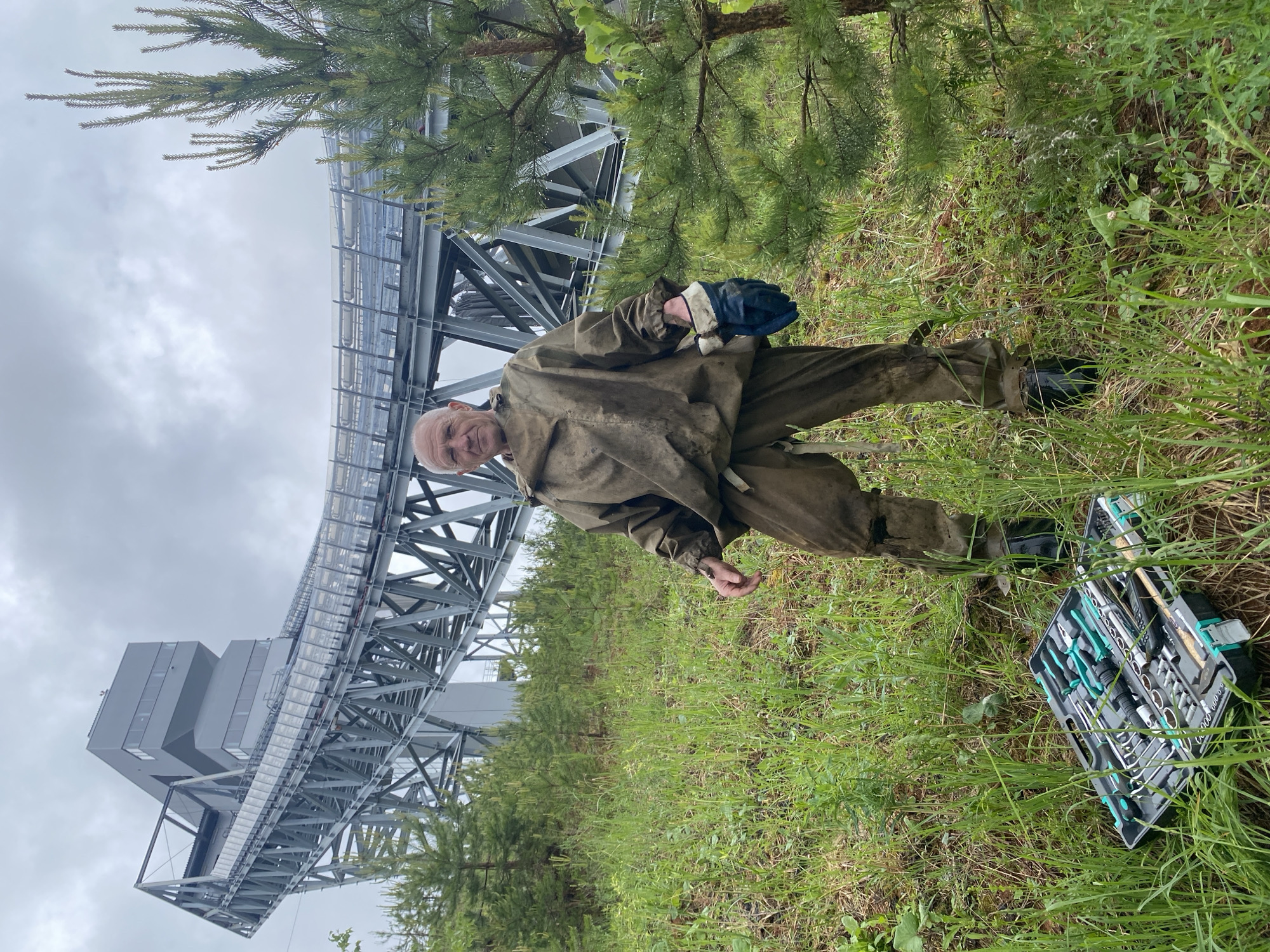 The image depicts an elderly man standing in a grassy field adjacent to a large, elevated structure, possibly a conveyor belt or a similar industrial installation. The location is near Прикамский, Russia, as indicated by the context.
The man is wearing what appears to be a weathered protective suit and work gloves. He is positioned in the foreground, facing the camera with a slight smile. To his right, on the ground, lies a toolbox filled with various hand tools.
The field is covered with green grass and small trees. The elevated structure is made of steel beams and supports a covered walkway. The sky is overcast.
