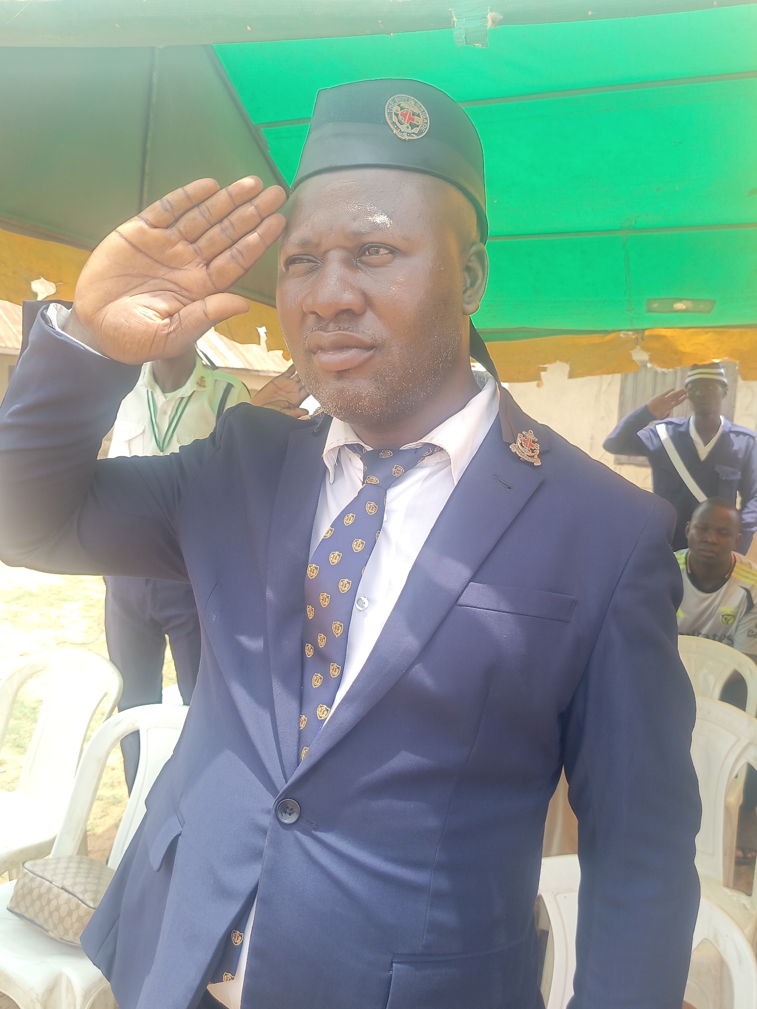 The image shows a man in Akwanga, Nigeria, wearing a blue suit, a white shirt, and a tie with a shield pattern. He also wears a dark cap with a circular emblem. He is giving a salute. In the background, other people are visible, one wearing a blue uniform and also saluting. White plastic chairs are scattered around the scene. A light-colored bag sits on a chair to the left. There is a green awning overhead.